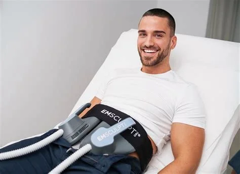 Man lying on a hospital bed with medical equipment strapped to his abdomen, smiling.