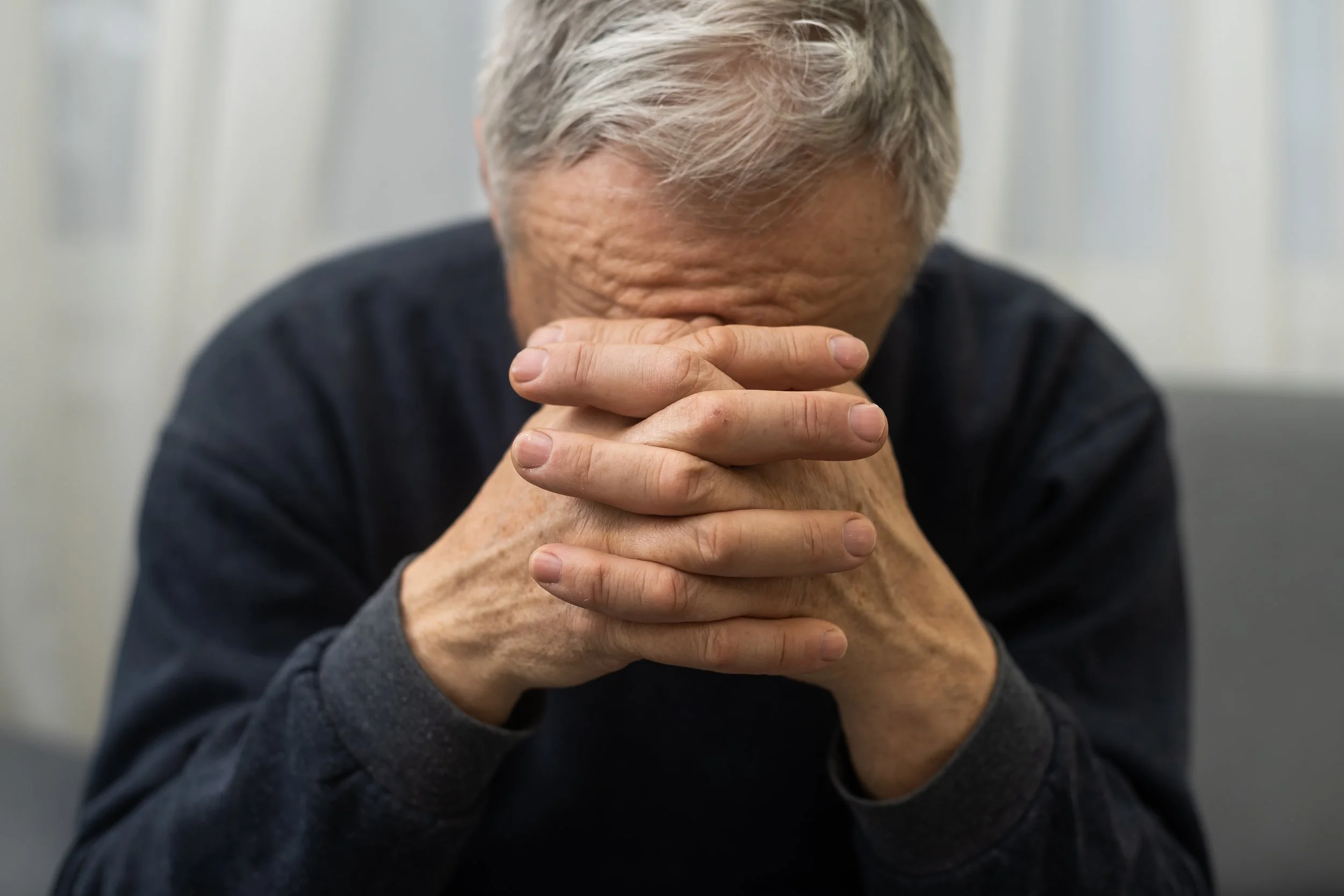 An older man with gray hair, wearing a dark shirt, has his head bowed and his hands clasped together, resting on his forehead.