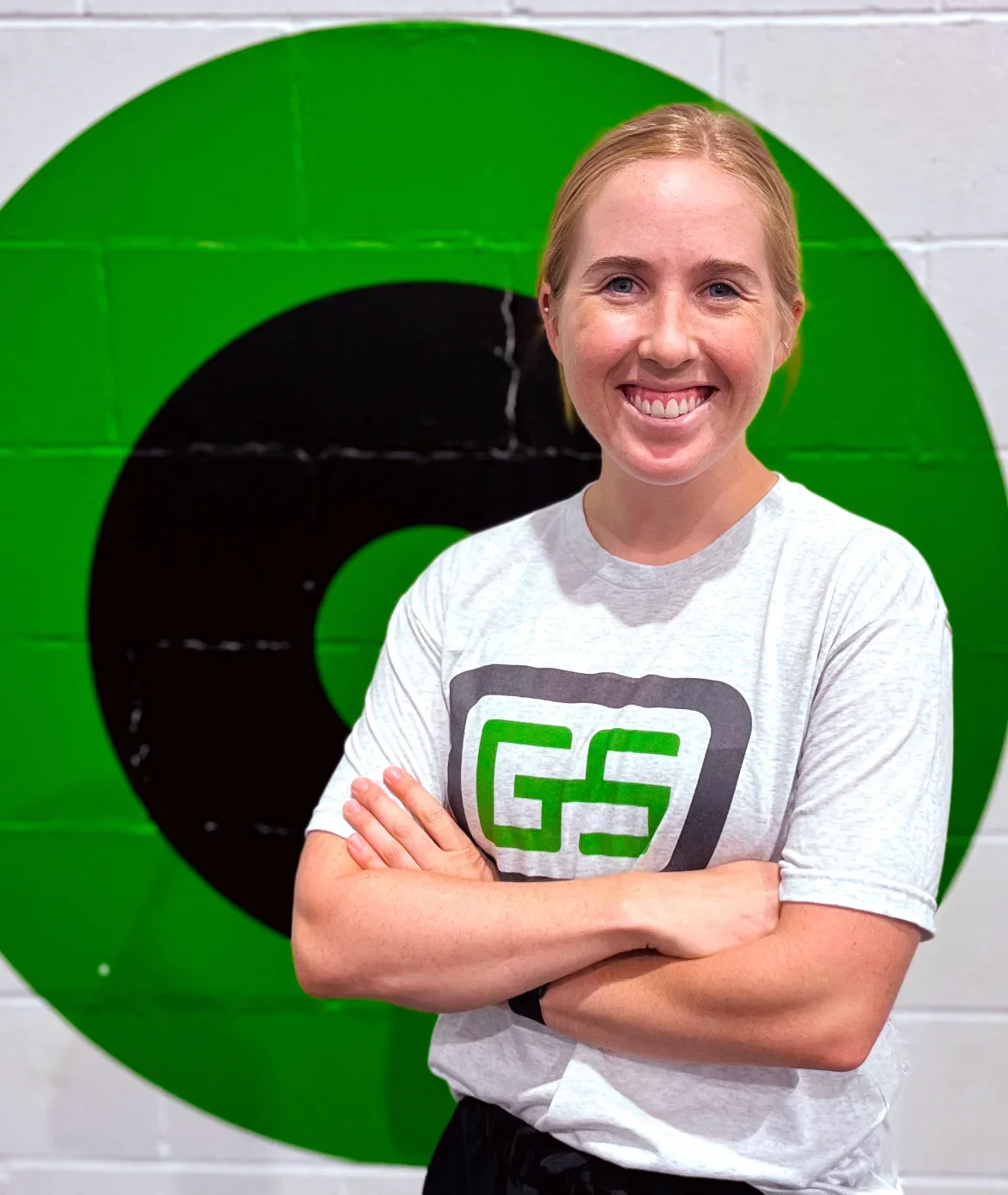 A smiling woman with blonde hair and crossed arms standing in front of a large green and black logo on a white brick wall. She is wearing a white T-shirt with a black and green logo that says 'GS'.