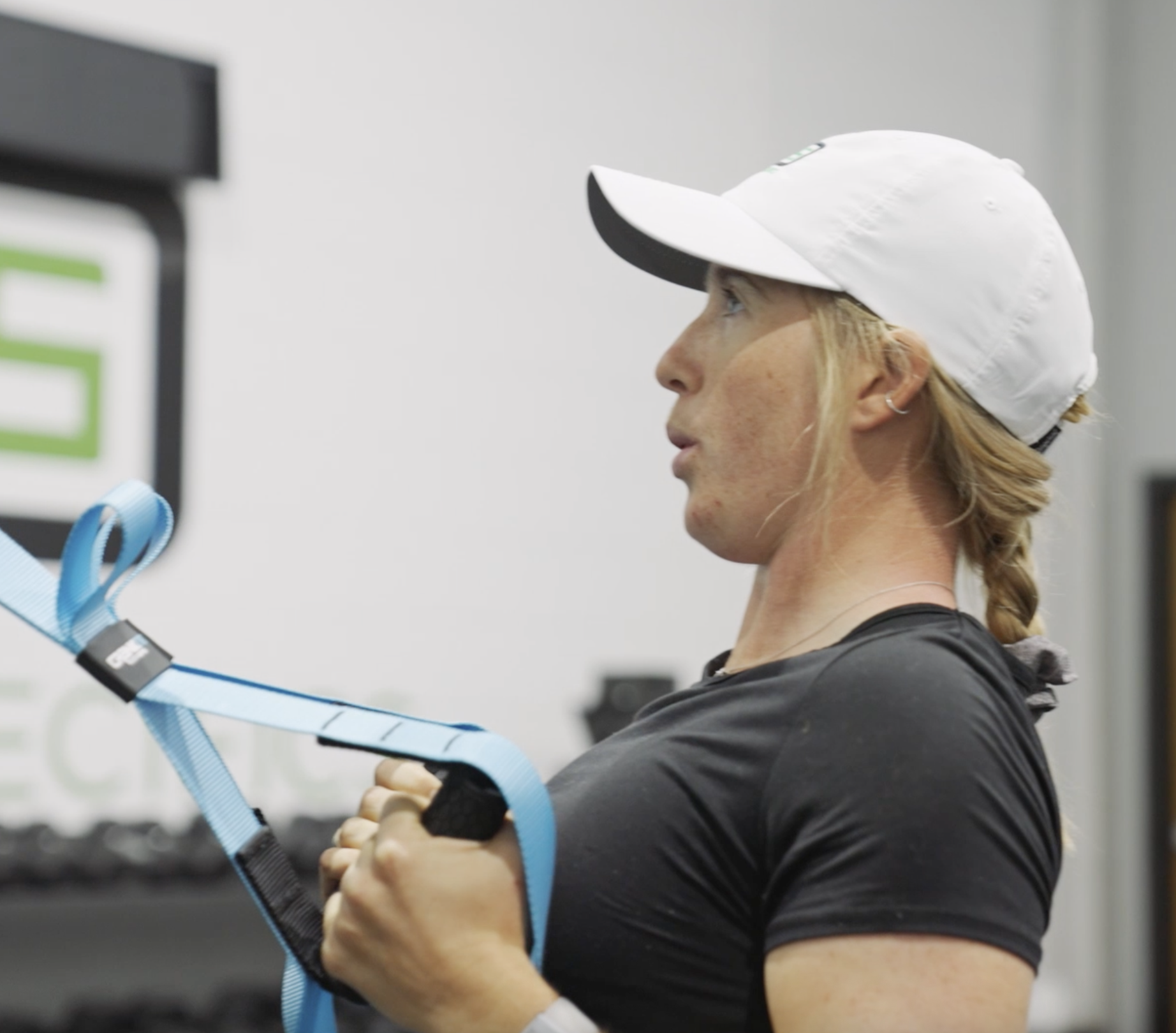 A woman wearing a white cap and black athletic shirt holds blue resistance bands during a workout.