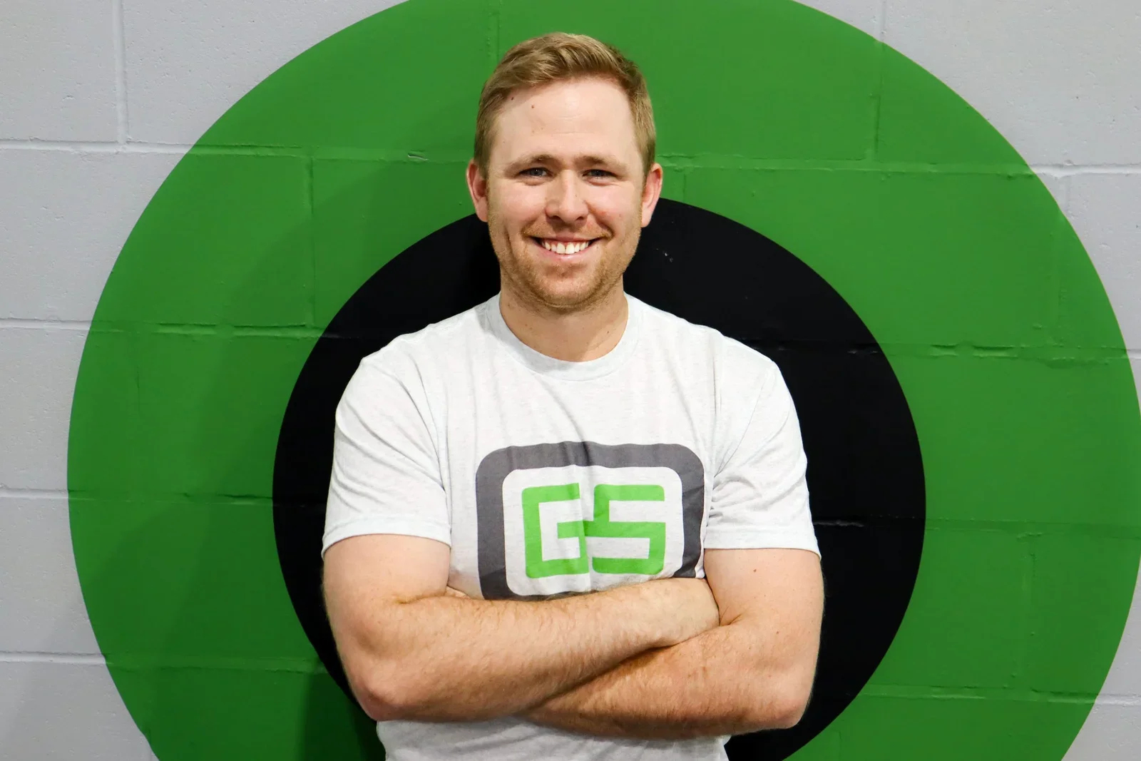 A smiling man with short blond hair and a beard standing with his arms crossed in front of a wall painted with a large target symbol in green, black, and white. He wears a white T-shirt with a logo that has the letters 'GS' in green and black.