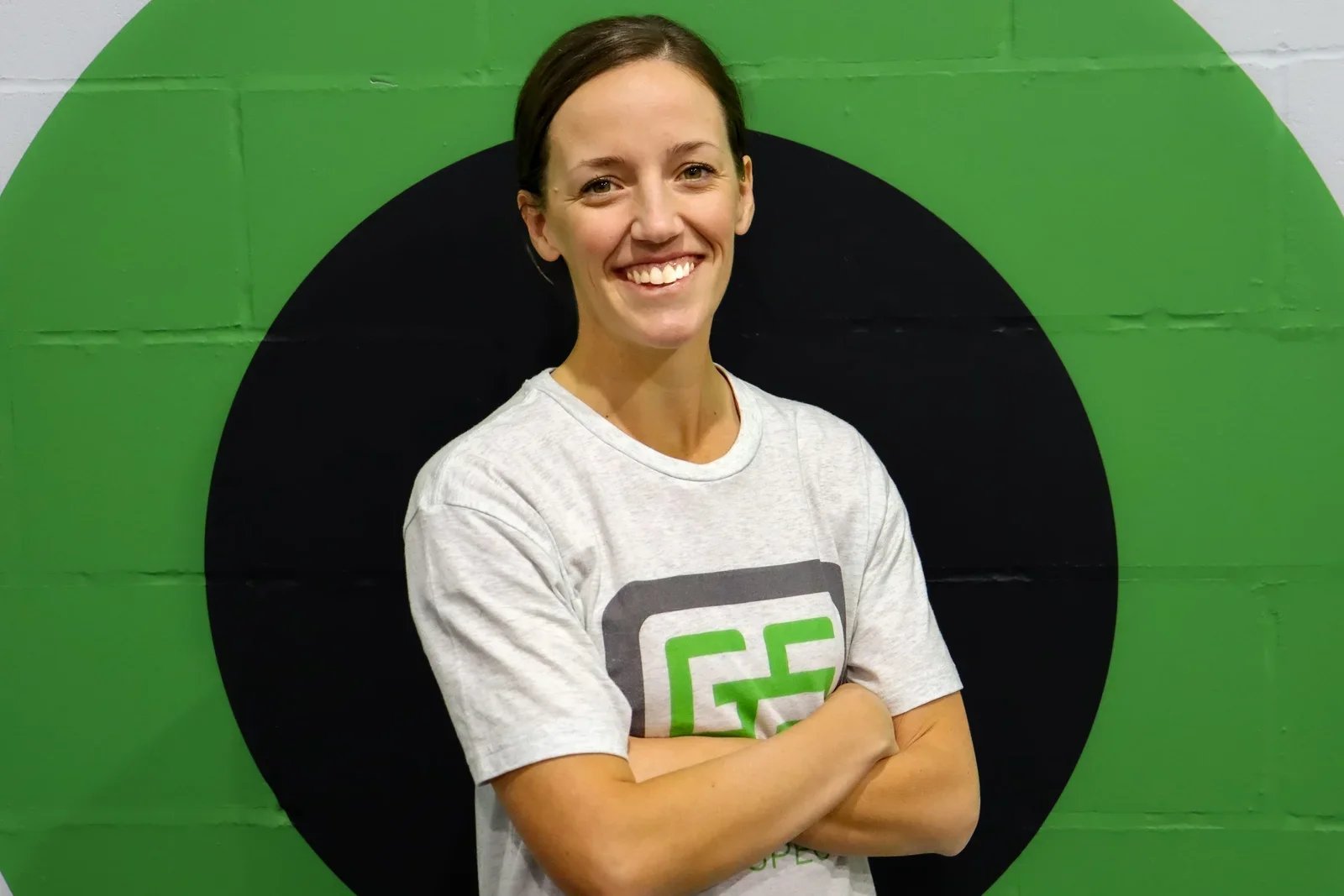 A woman smiling with arms crossed, standing in front of a green wall with a large black circle.