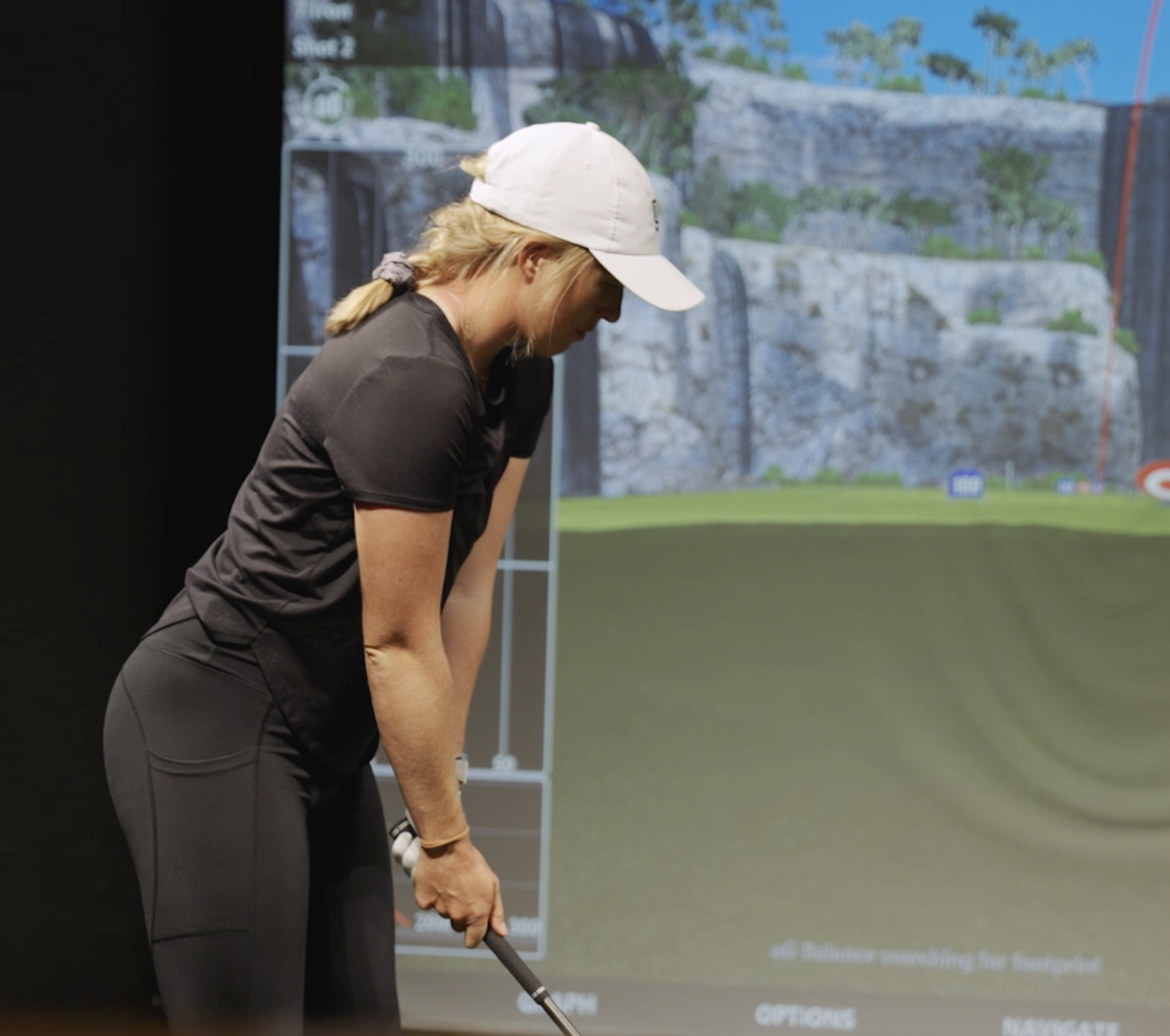 A woman wearing a white cap and black athletic clothing is preparing to hit a golf ball on an indoor simulator with a large screen displaying a golf course.