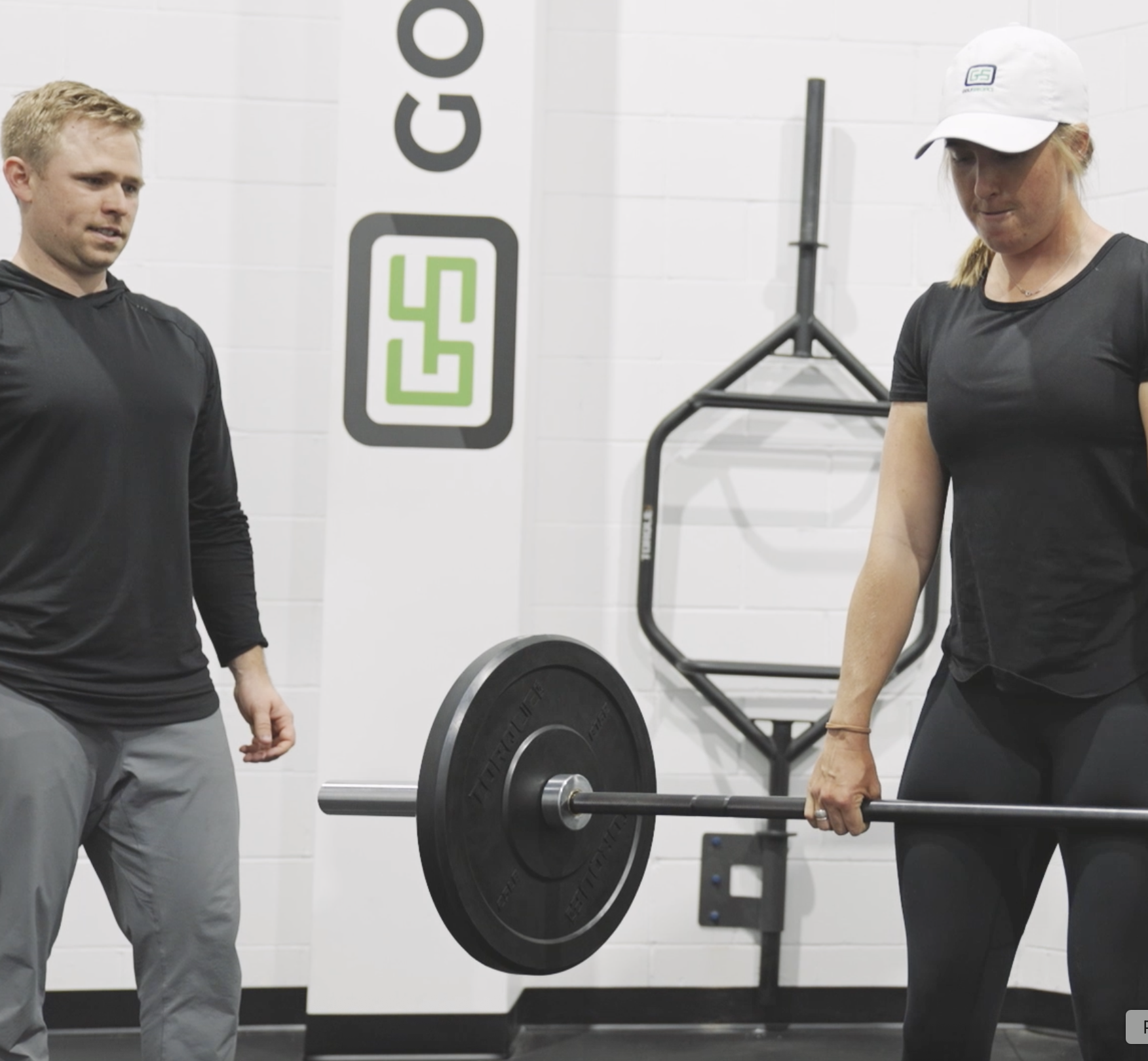 A woman in black workout clothes is performing a deadlift with a barbell, while a man in black and gray workout clothes watches in a gym with white tiled walls and gym equipment in the background.