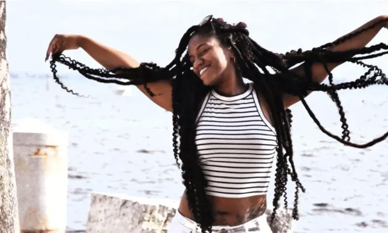 A smiling woman with long braids and a striped sleeveless crop top stands by the water with arms outstretched.