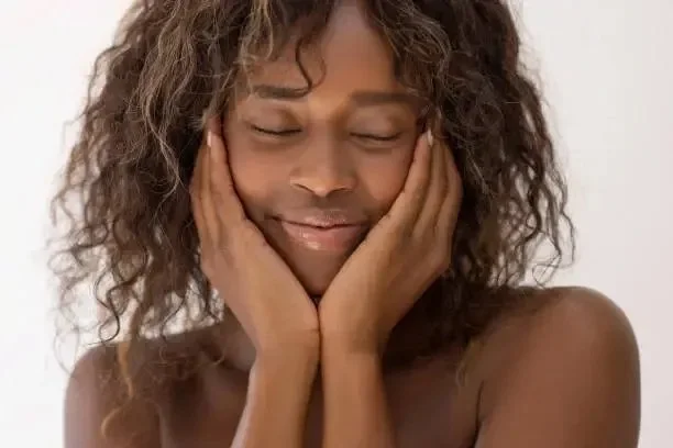 A woman with curly hair gently touching her face with closed eyes and a peaceful smile.
