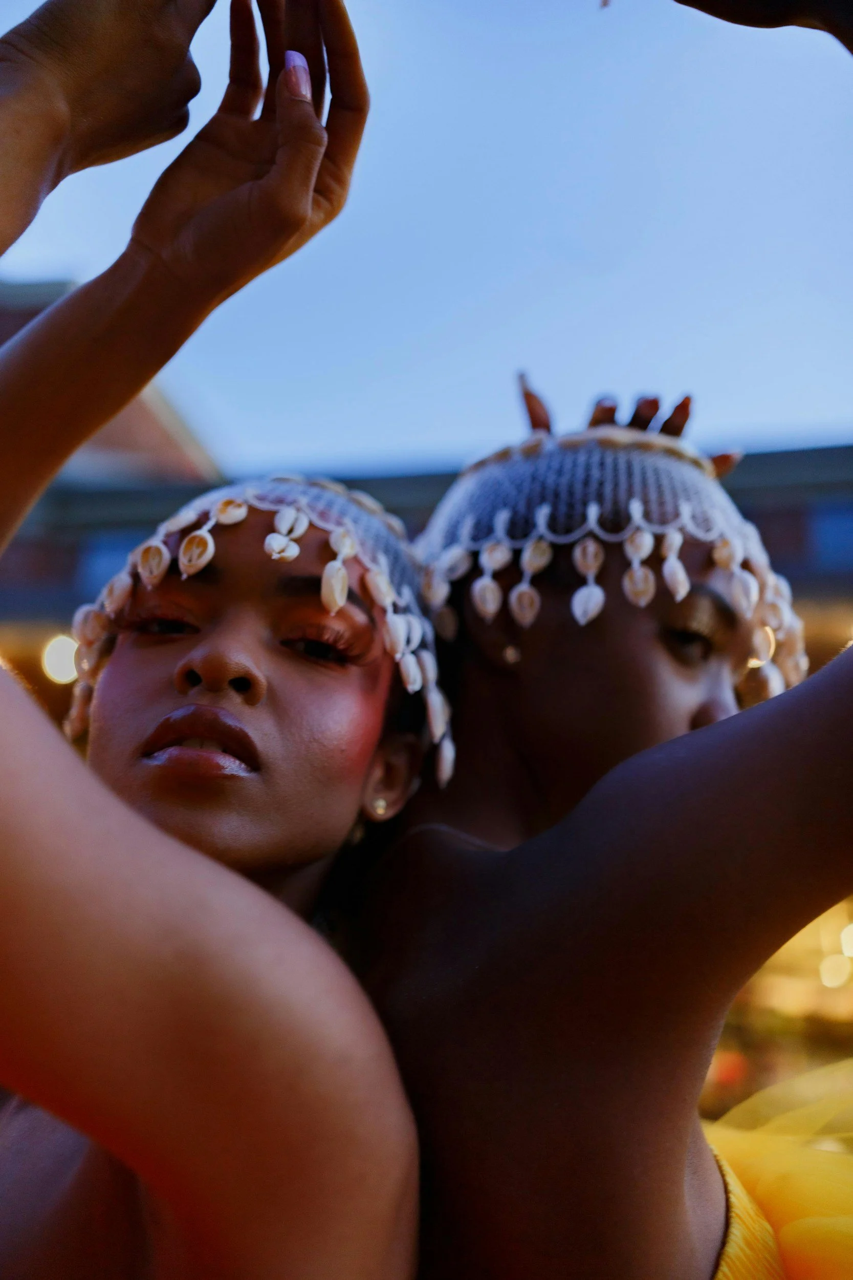 Two women with hair adorned in seashells, posing closely together outdoors during dusk or evening.