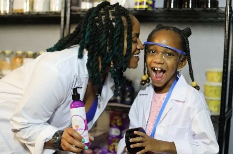 A woman and a young girl wearing lab coats and safety goggles are sharing a joyful moment in a store aisle with shelves of jars and containers behind them. The girl is smiling widely and holding a device, while the woman is holding a bottle of lotion.