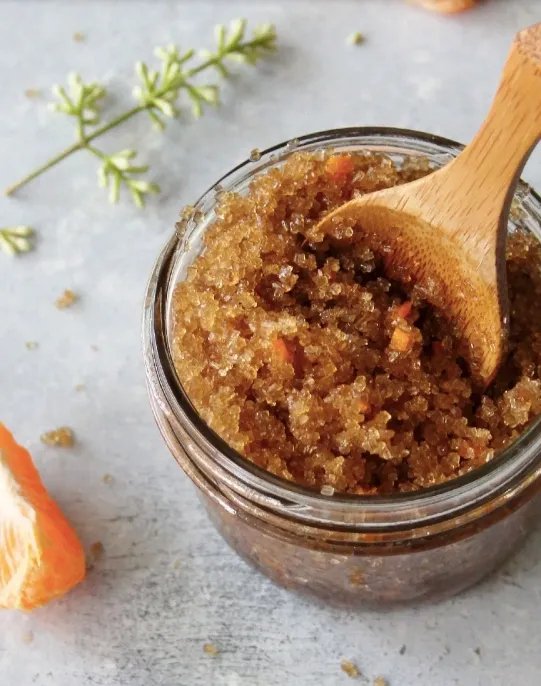 Close-up of a jar of chunky brown homemade fruit jam or preserves with a wooden spoon inside, on a light surface with a slice of orange and some green herb in the background.