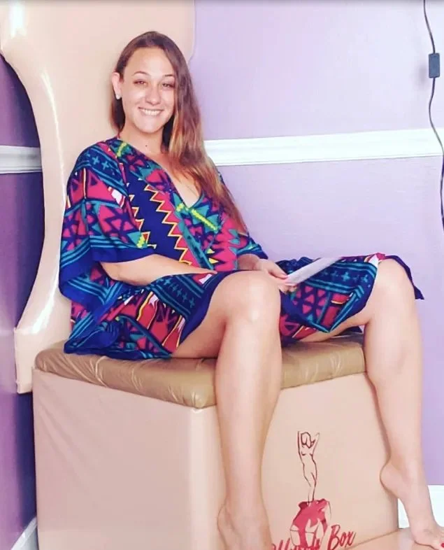 A woman with long hair and a bright, multicolored dress is sitting on a padded medical examination table. She is smiling and holding a piece of paper, with her legs extended and feet resting on the table. The wall behind her is painted purple with a white chair rail, and there are visible wires on the wall.