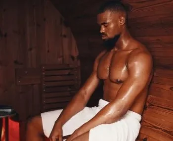 A shirtless man with a towel around his waist sitting against a wooden wall.