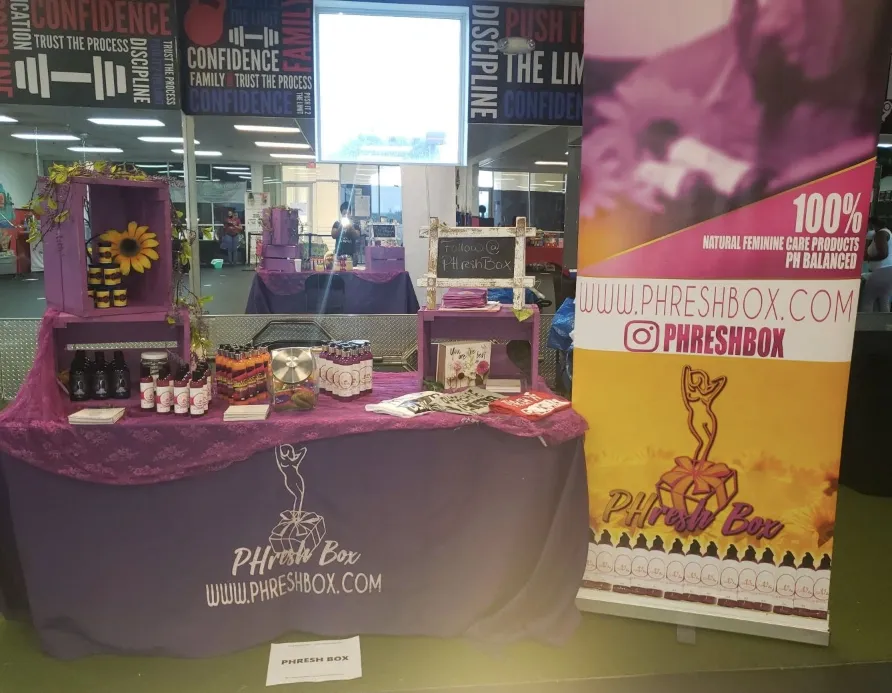 Display of health and wellness products at a booth, decorated with purple cloth, featuring bottles, jars, and a small chalkboard sign, with a promotional banner on the side promoting PHresh Box for natural feminine care products.