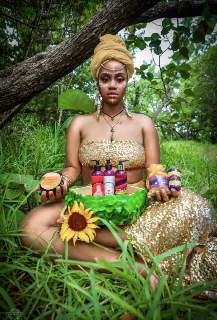 Young woman with a tan headwrap and gold sequin dress sitting in grass under a tree, holding bottles and a sunflower, surrounded by various beauty products.