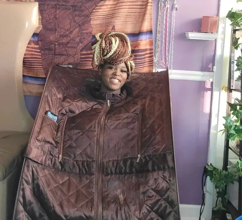 Woman smiling inside a portable blanket sleeping bag with a zipper, sitting on a chair in a room with purple walls, a map of Africa, and decorative curtains.