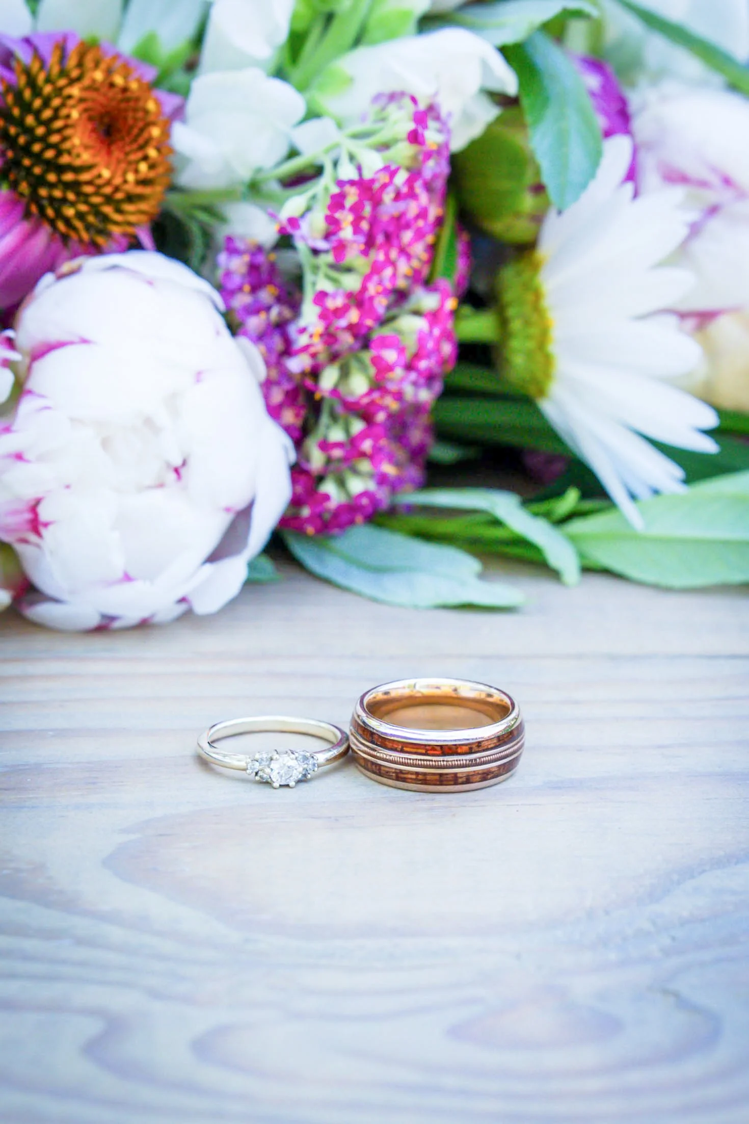 A bouquet of colorful flowers, including white peonies, and purple and pink blossoms, with two rings, one with a diamond and the other with a textured pattern, on a light wooden surface.