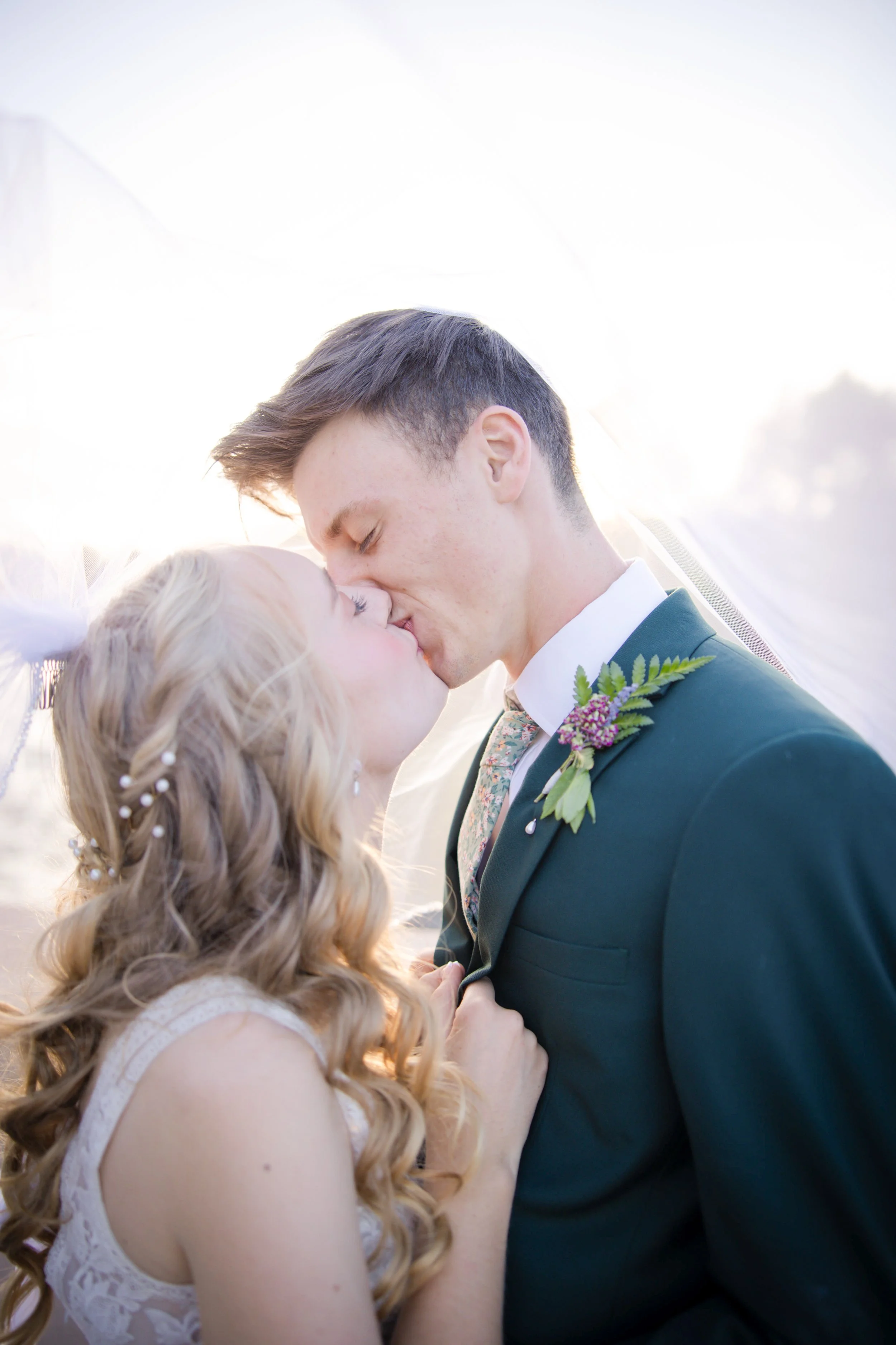 A bride and groom share a kiss, standing close under a white veil, during their wedding, with bright sunlight behind them.