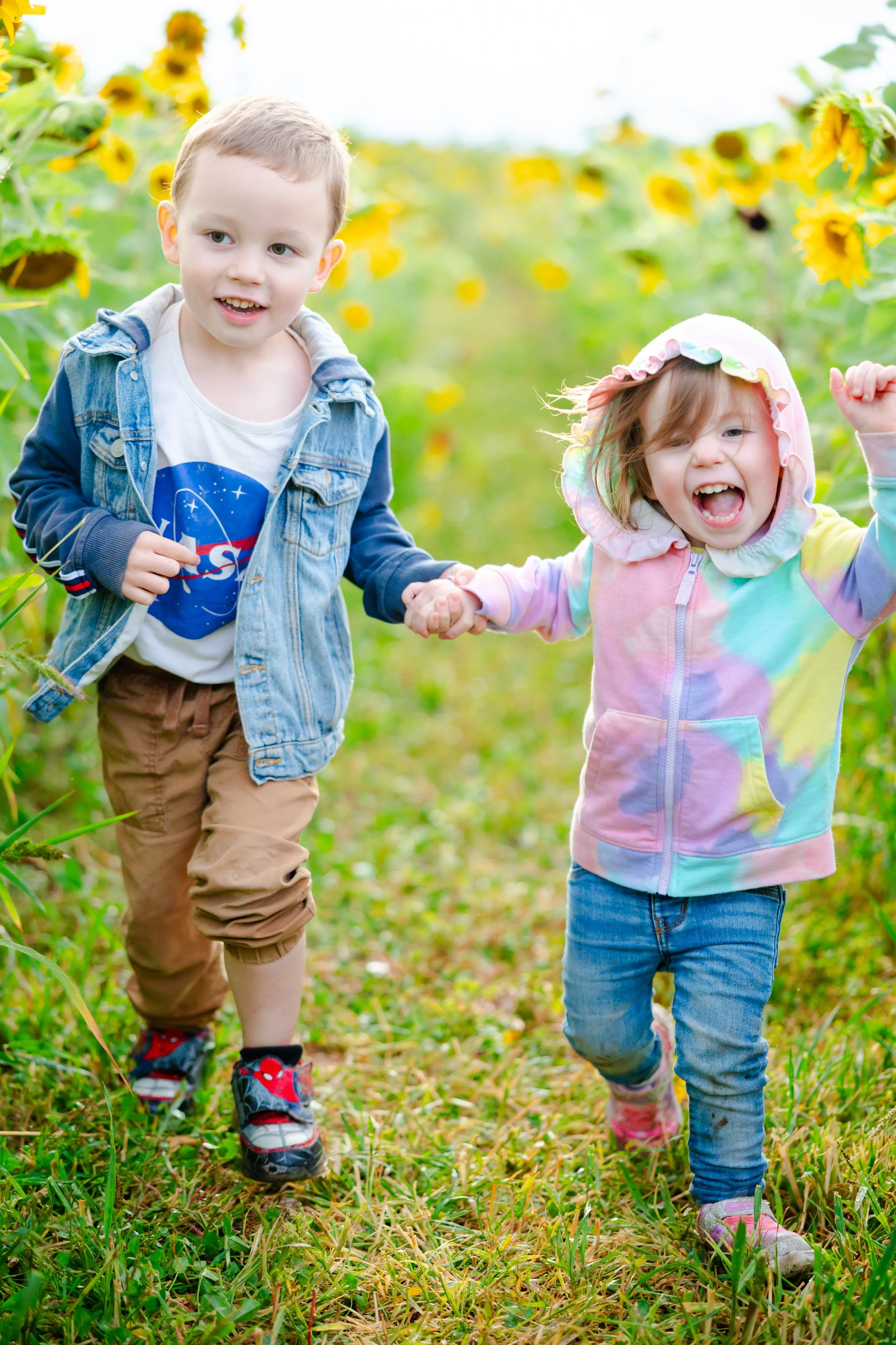 Two children holding hands and running through a sunflower field, smiling and enjoying outdoors.