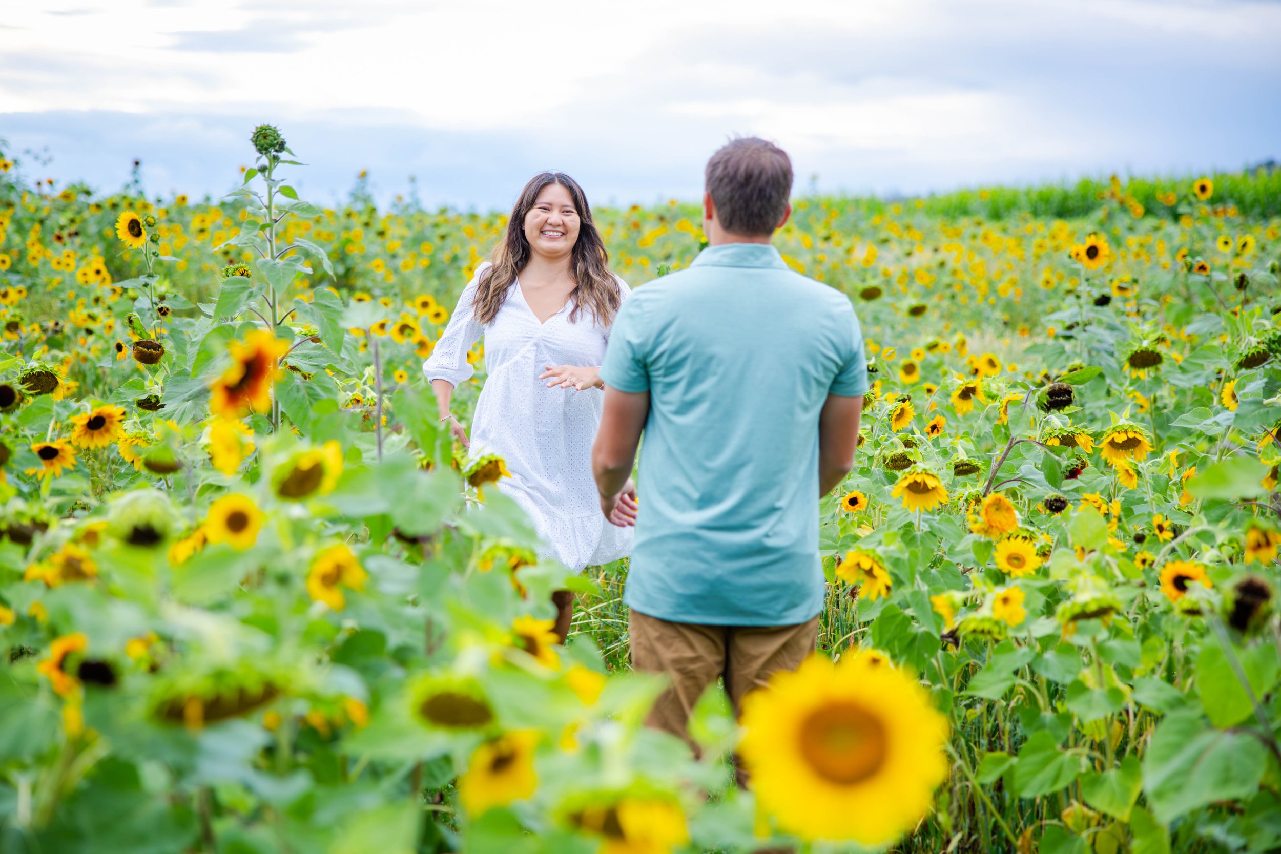 A man and woman standing in a sunflower field, smiling and talking, with a cloudy sky above.