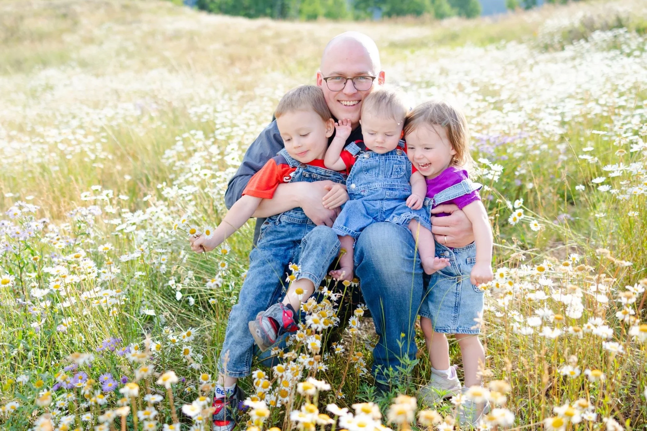 A man with glasses and a bald head smiling in a field of daisies, holding three young children, all dressed in casual clothes, surrounded by blooming daisies on a sunny day.