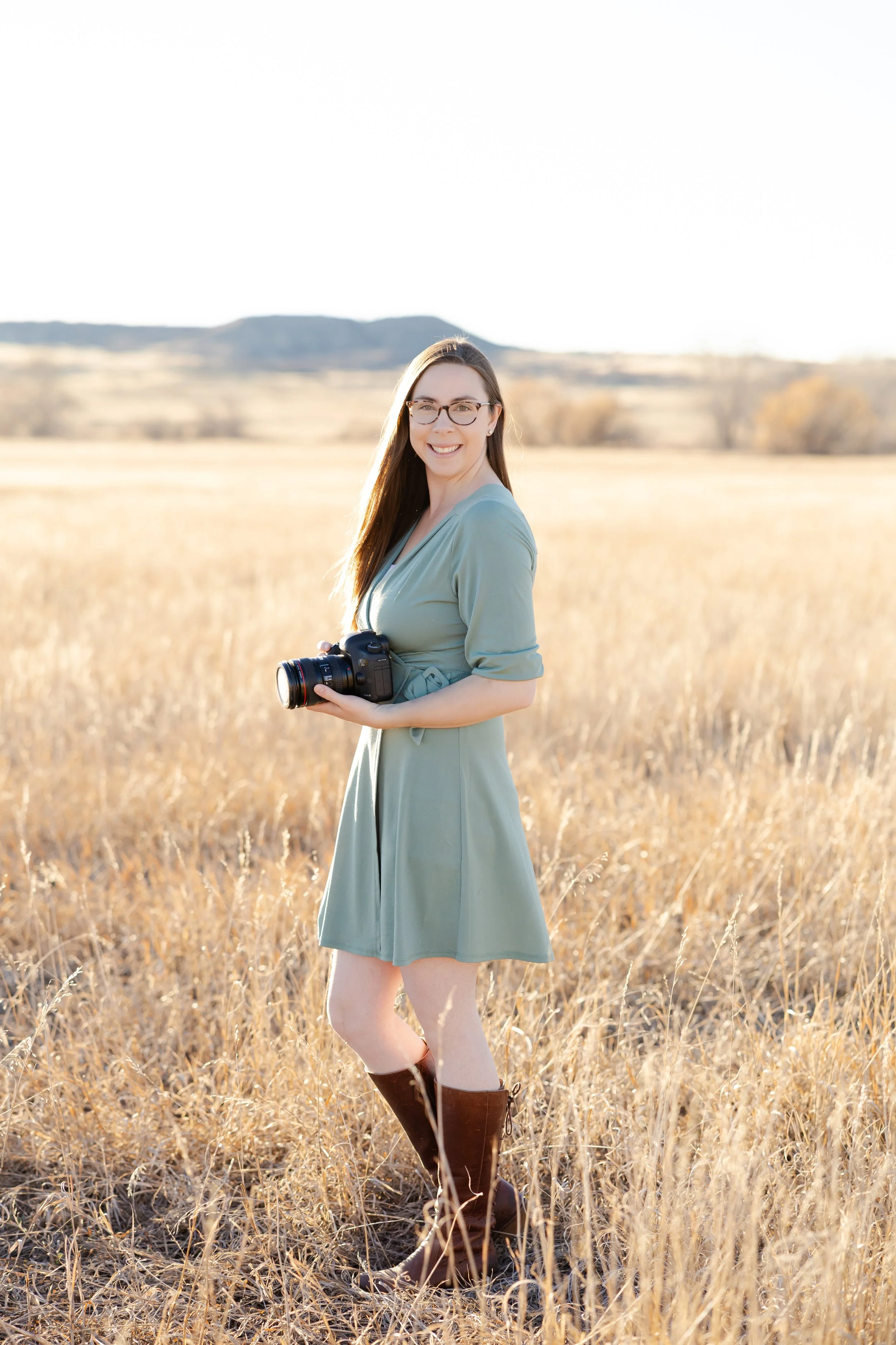 A woman standing in a field of tall, golden grass, holding a camera, smiling, wearing a green dress and brown knee-high boots, with mountains in the background during the daytime.