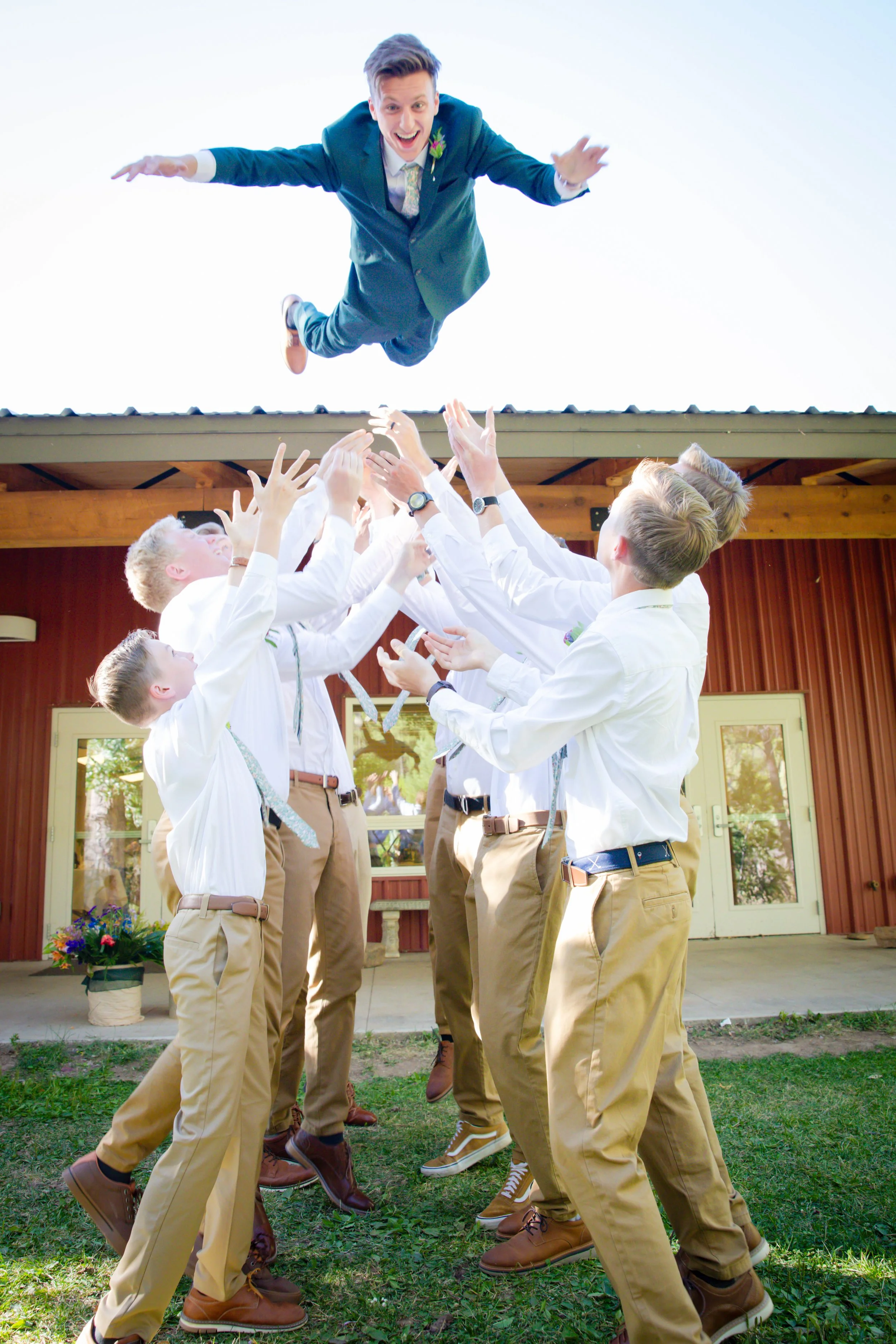 A group of young men in dress shirts and khakis celebrating outdoors as they throw a man in a blue suit into the air during a wedding celebration.