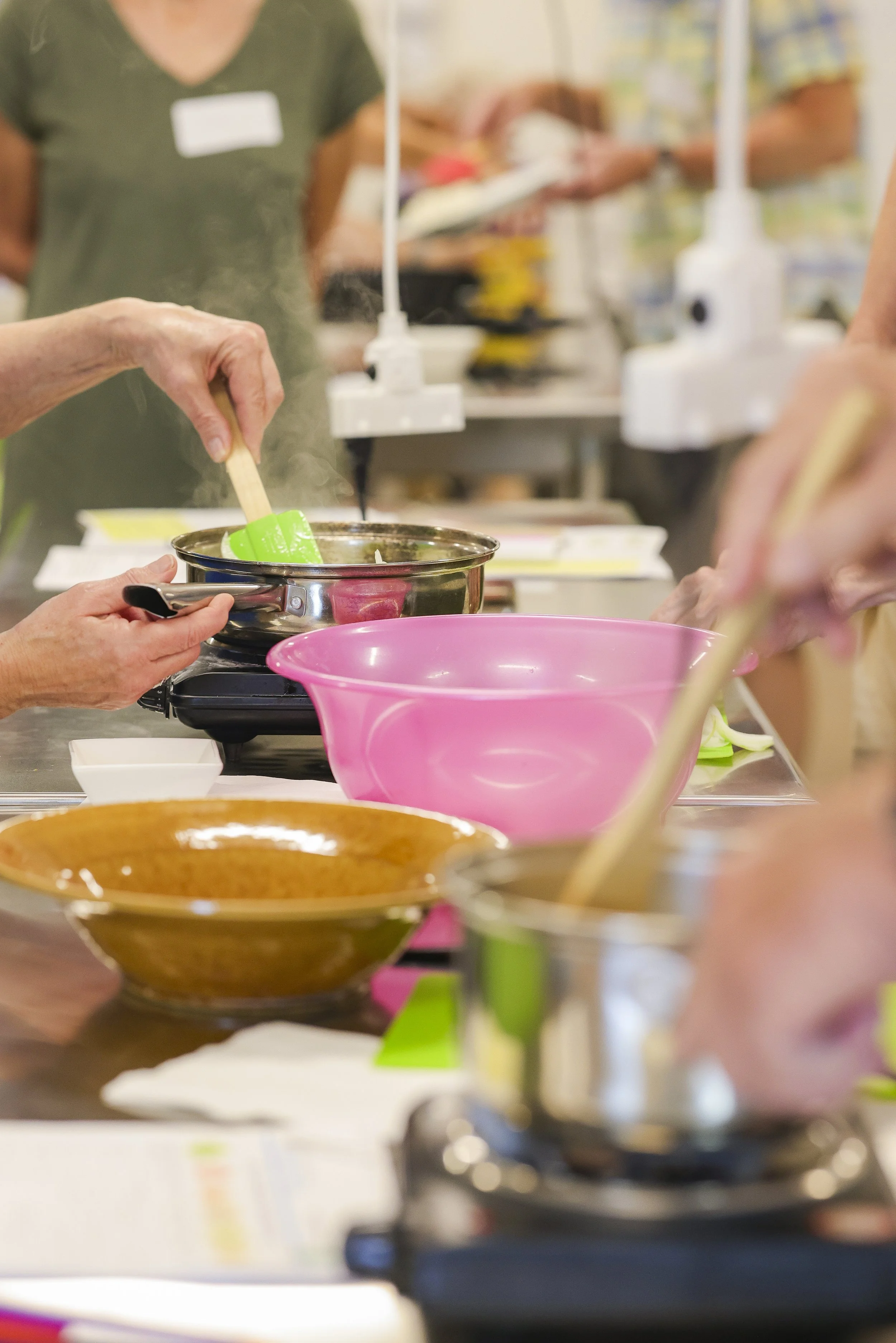 People participating in a cooking class, stirring and preparing food at a table filled with bowls, utensils, and a stovetop.