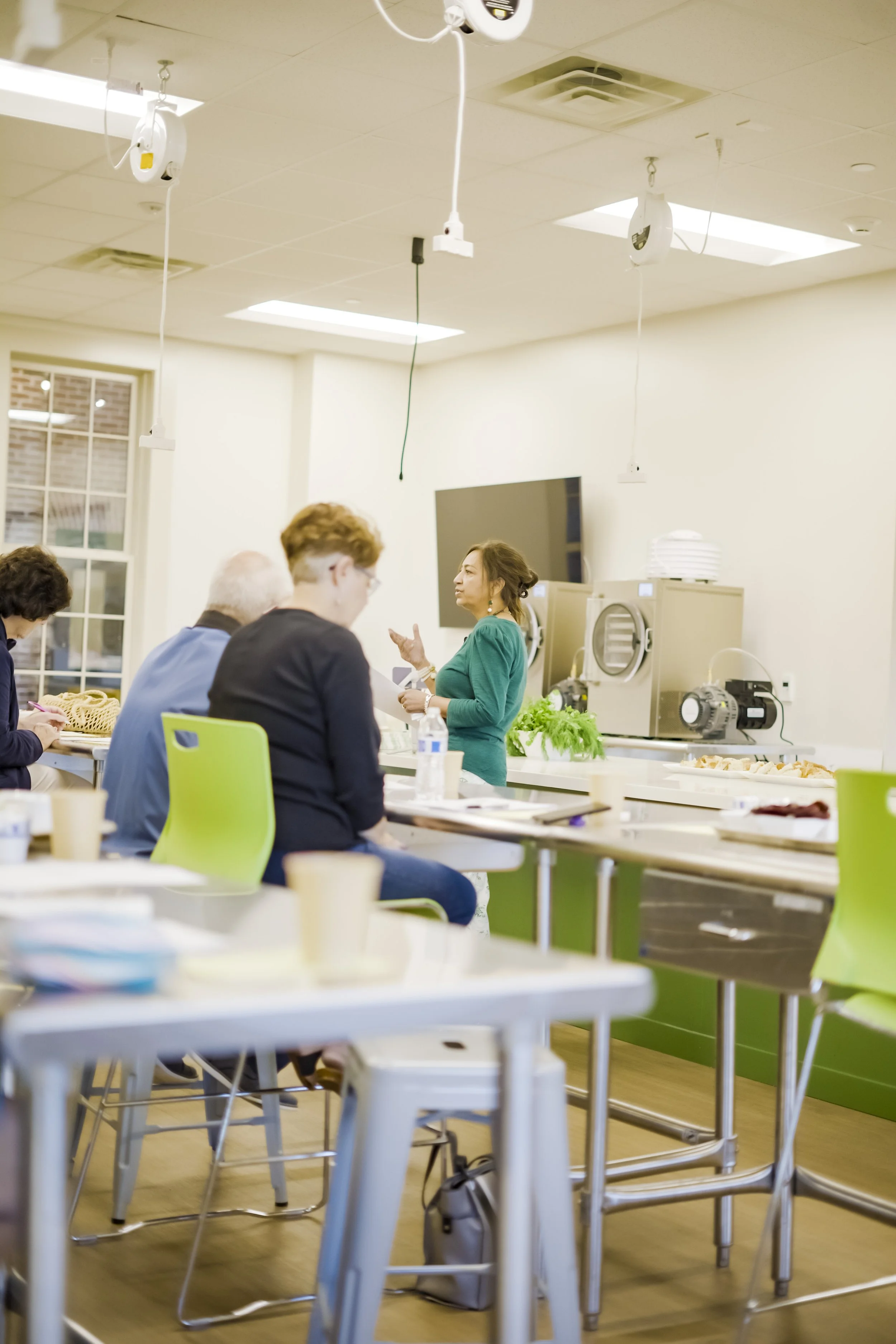 A woman in a teal dress leading a workshop or presentation in a well-lit room with green chairs, tables with papers and cups, and some equipment in the background.