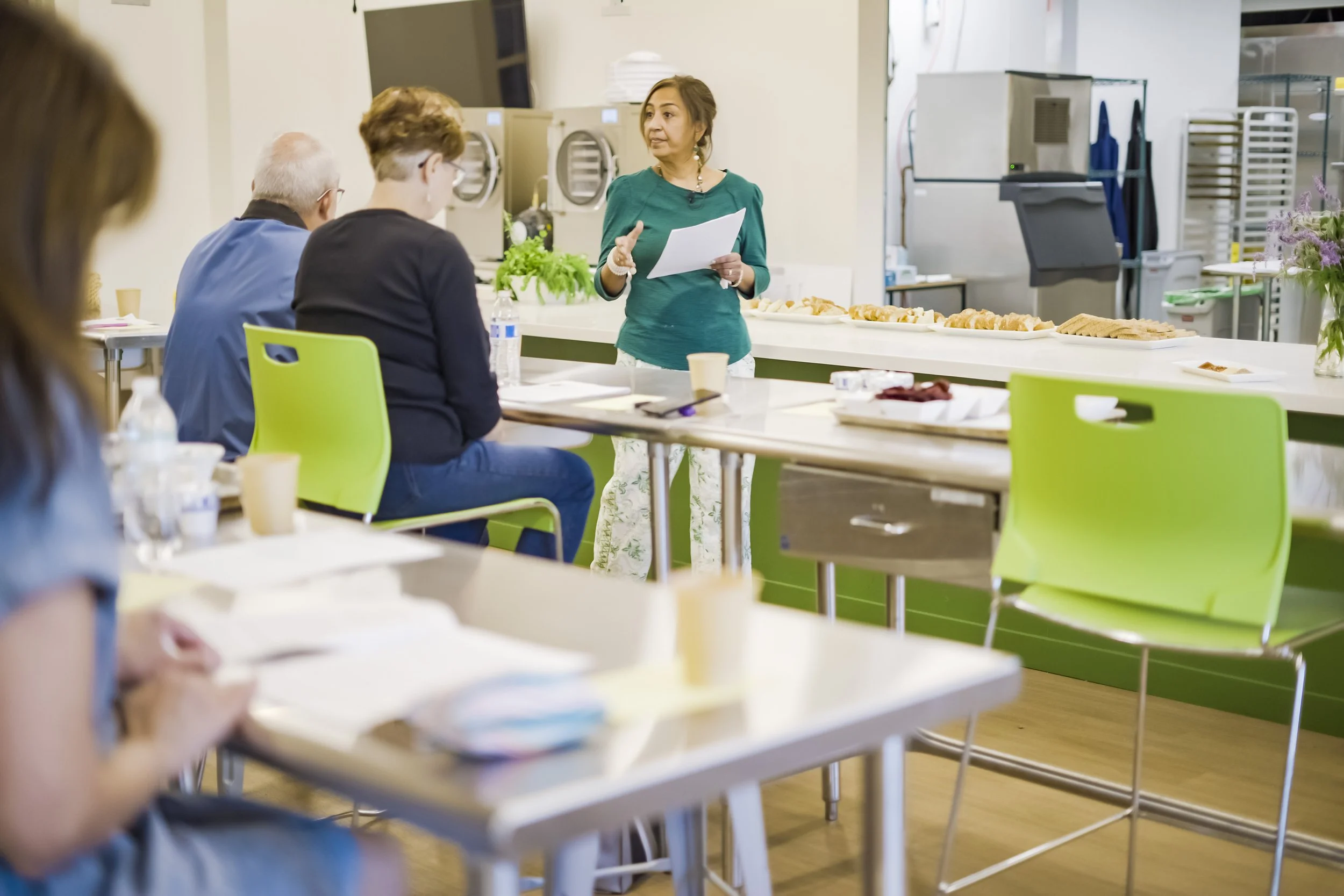 A woman in a teal top giving a presentation to a small group in a modern, well-lit kitchen or cafeteria. The group, seated at tables with papers and cups, is listening attentively. On the counter behind her, there are snacks and a refrigerator in the