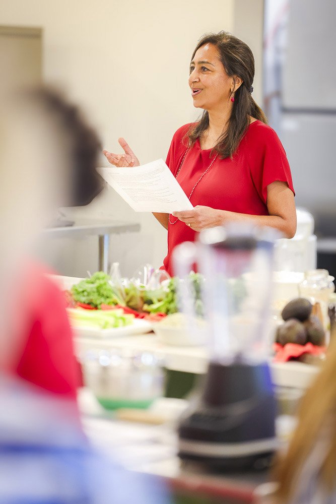 A woman in a red shirt standing in front of a table with vegetables and kitchen items, speaking or giving instructions to others.