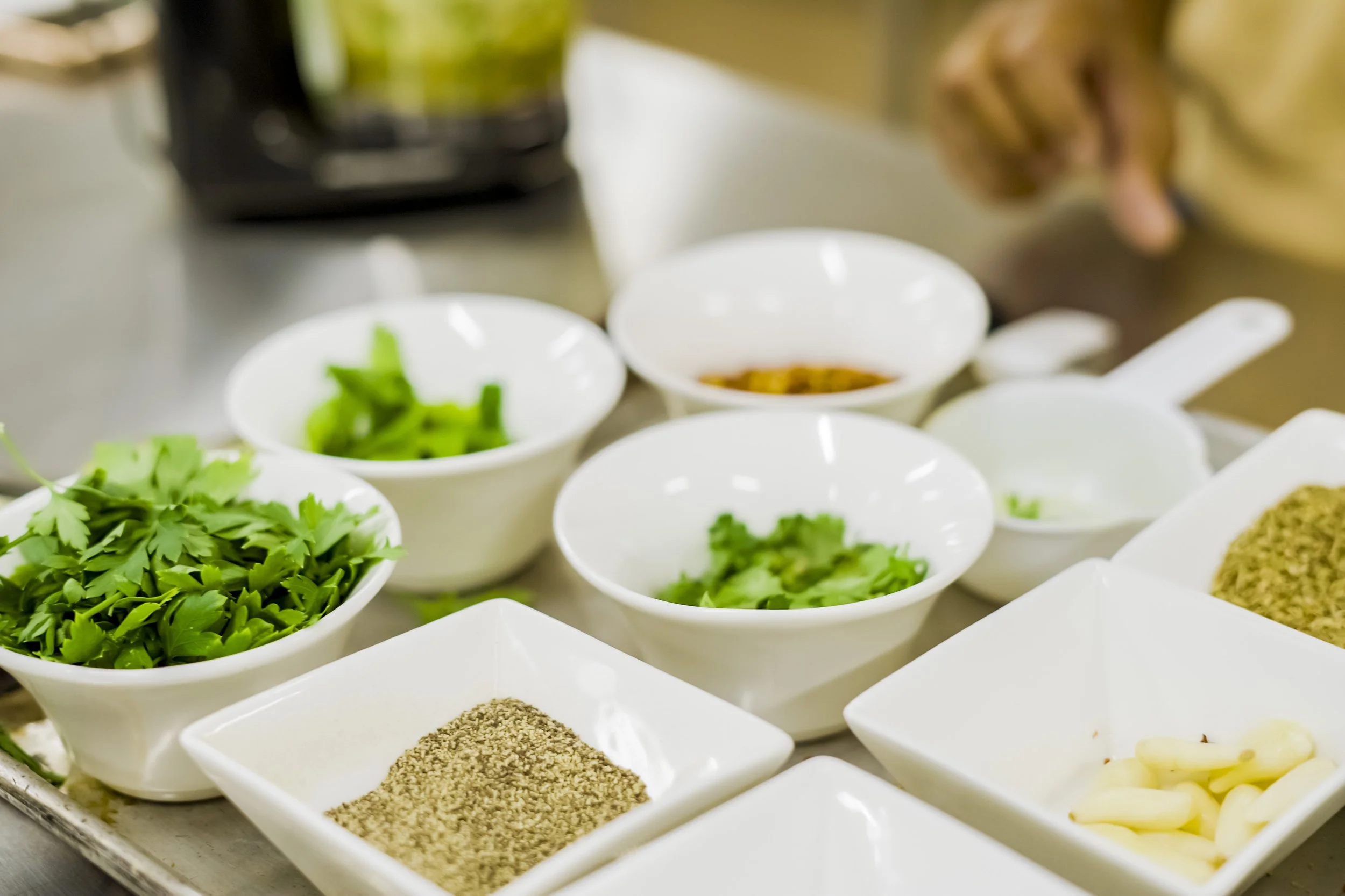 Various bowls and plates with chopped green herbs, ground spices, garlic cloves, and sauces on a kitchen counter.