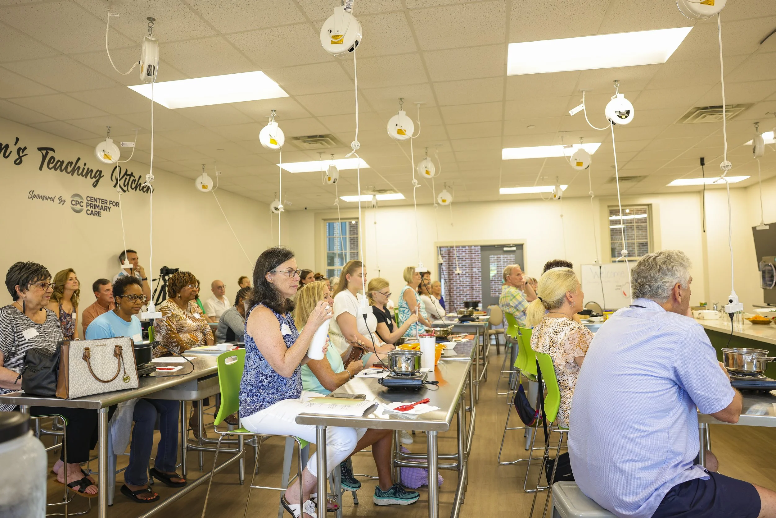 Group of people attending a cooking class in a bright room with hanging kitchen tools, some are taking notes, others are listening attentively