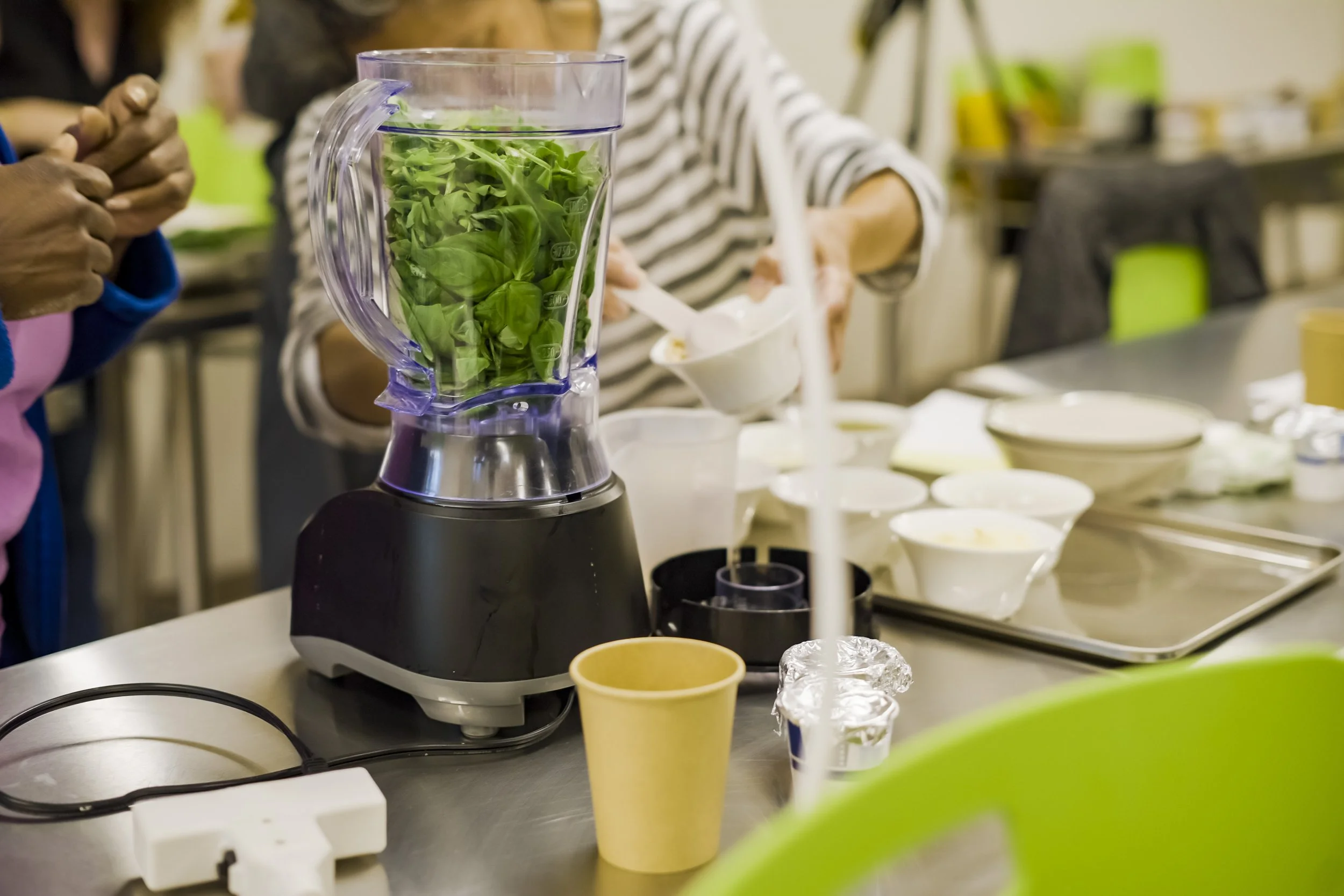 A kitchen or food prep area with a blender filled with leafy greens, surrounded by bowls, cups, and people preparing food.