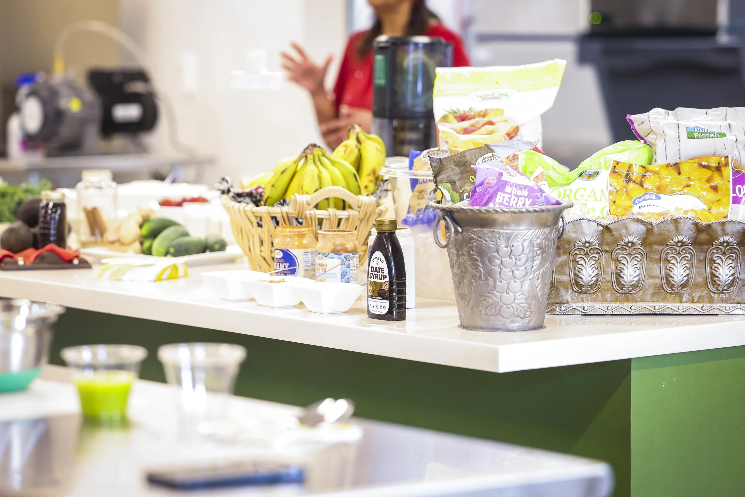 A kitchen counter filled with bananas, avocados, various packaged frozen fruit, and condiments. A person in a red shirt is in the background, slightly out of focus.