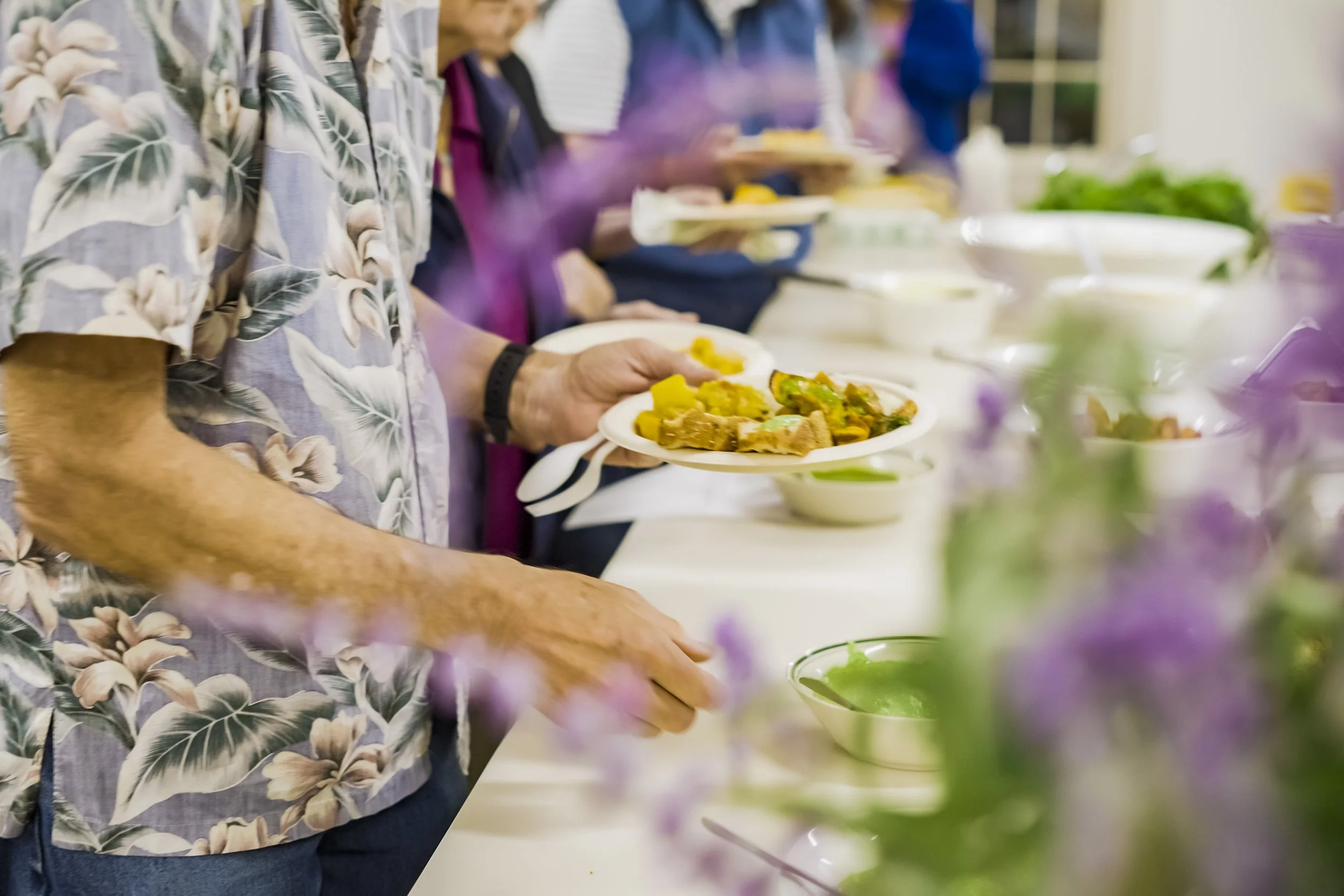 People standing in a line with plates of food at a buffet, featuring various dishes and green salads.