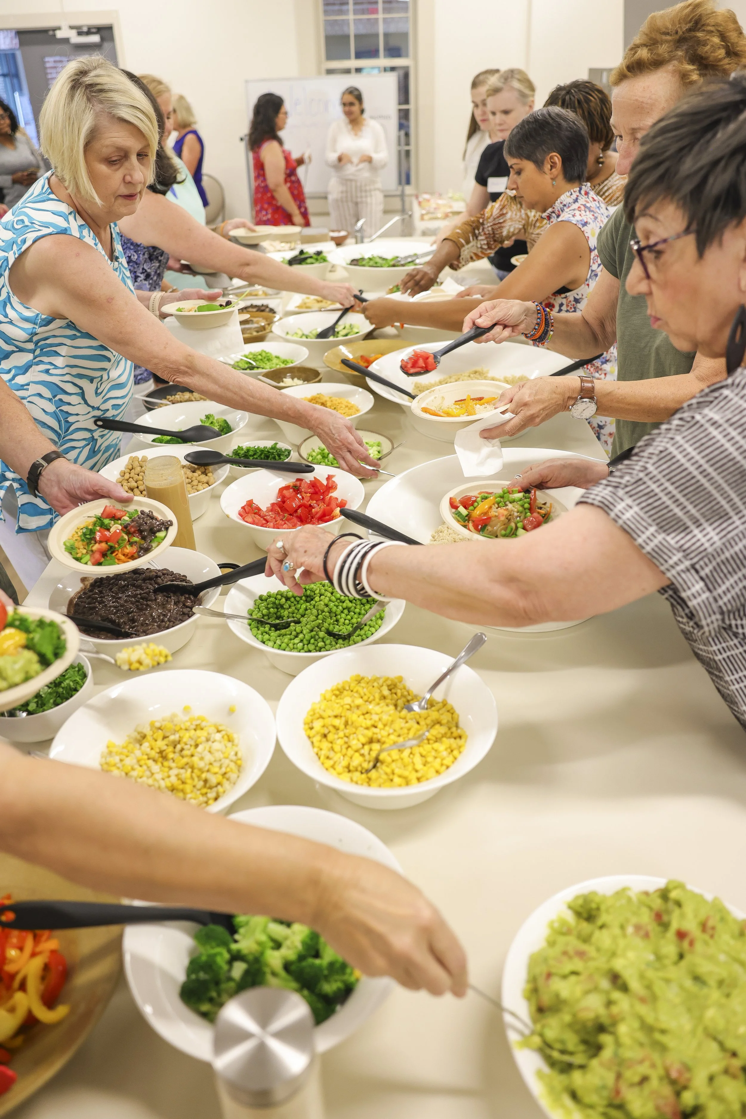 People serving themselves various salads and vegetables from a buffet table in a bright, indoor setting.