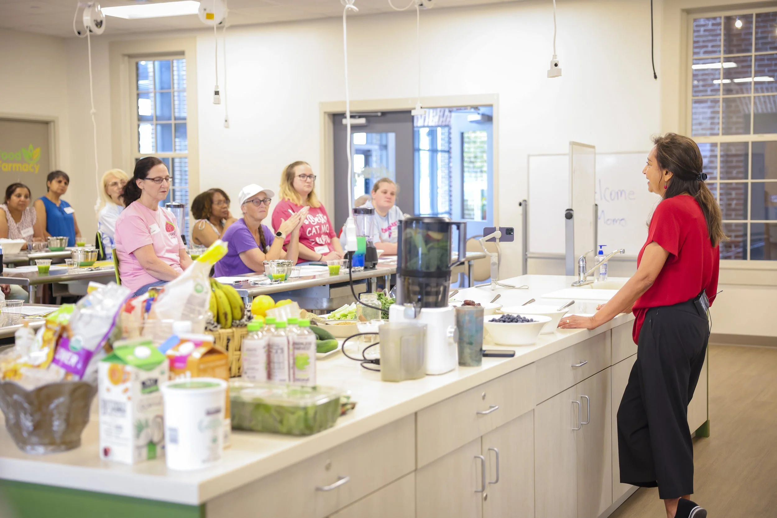 A woman in a red shirt gives a cooking demonstration to a group of women seated at a long kitchen counter. The counter is filled with ingredients and kitchen tools, and the room has large windows and modern decor.