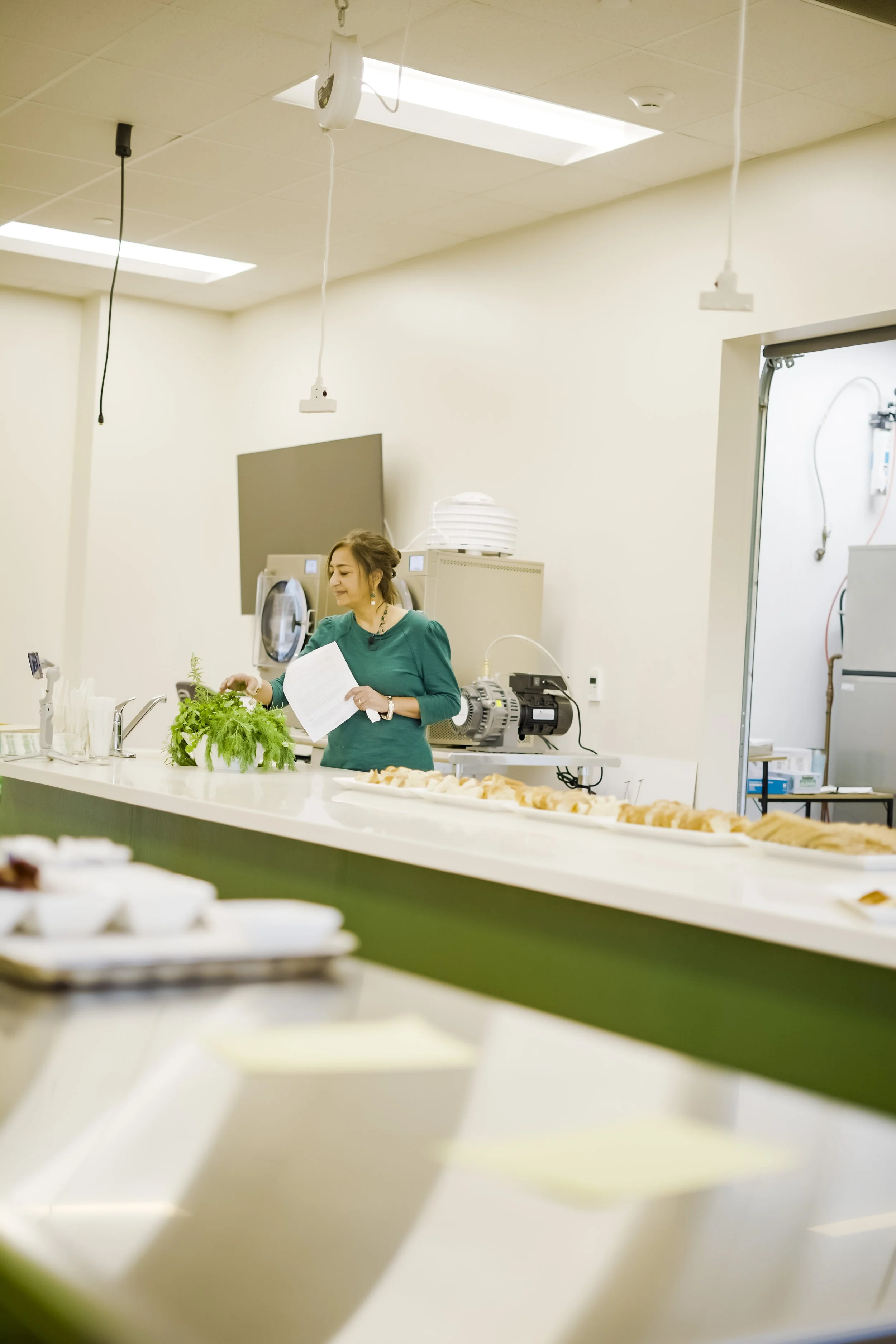 A woman in a green top preparing food or herbs in a kitchen or food service area, with trays of pastries or baked goods and various kitchen equipment around her.