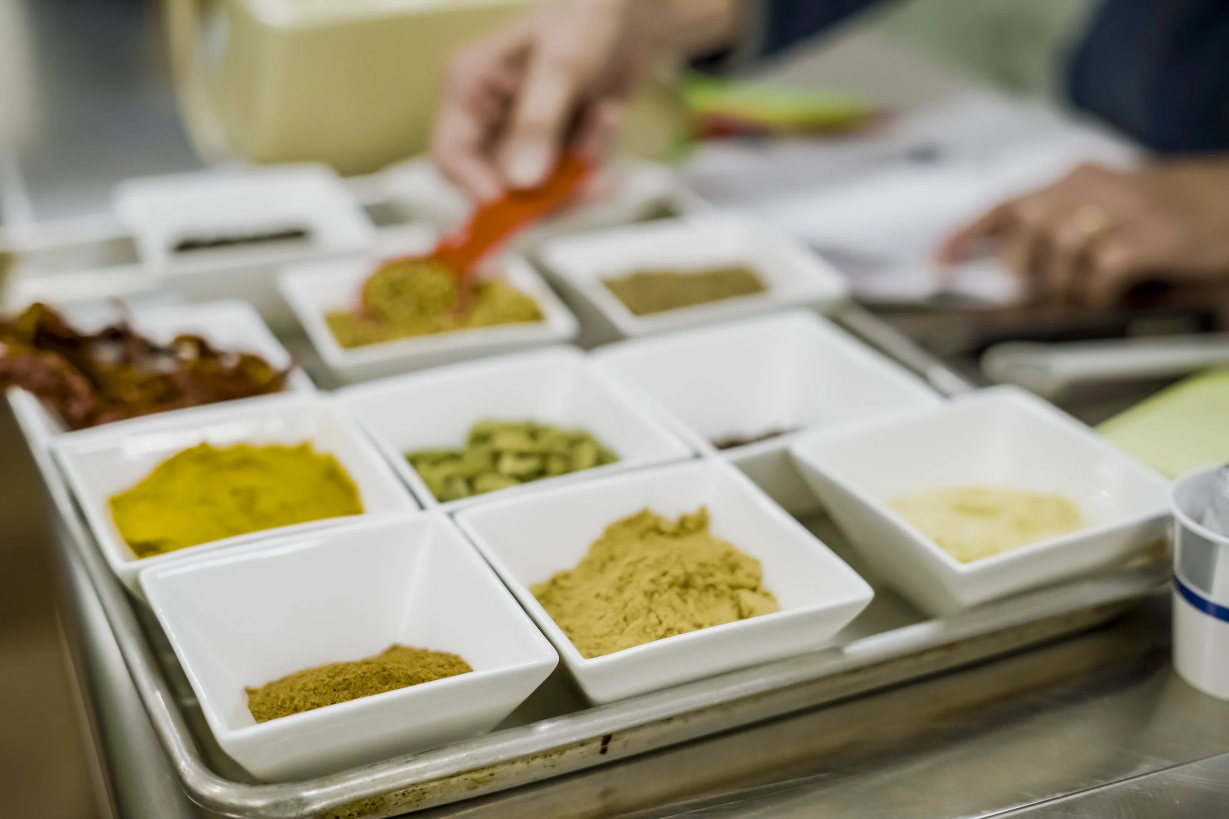 Assortment of spices in small white bowls on a stainless steel tray, with a hand adding a spice.