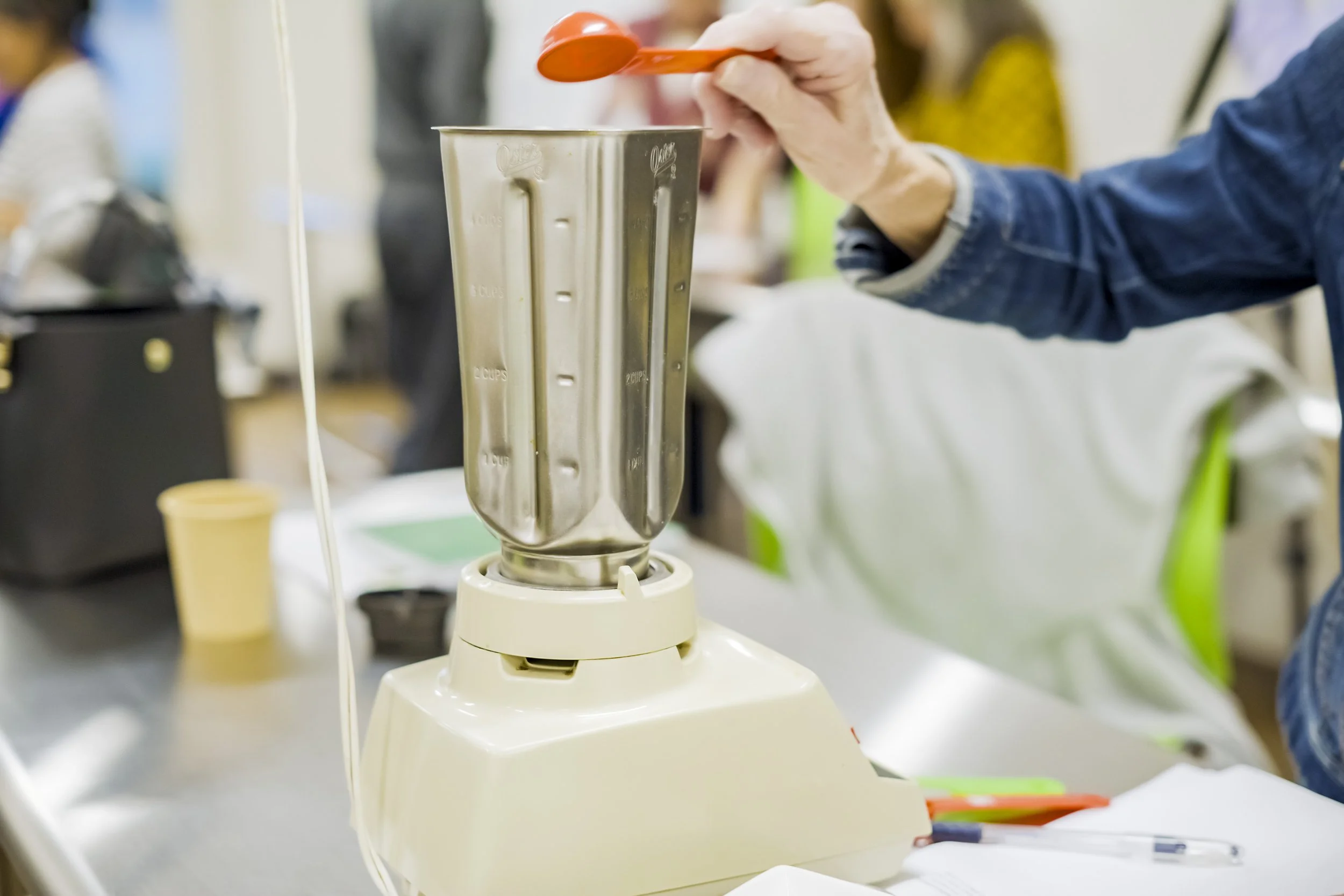 Person pouring ingredients into a blender in a kitchen or workspace with other people in the background.