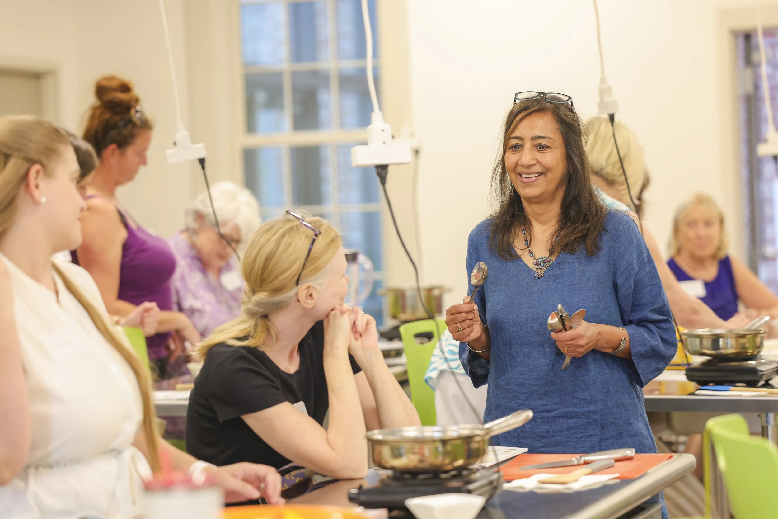 A woman teaching a cooking class to a group of women in a bright kitchen, holding cooking spoons and smiling.