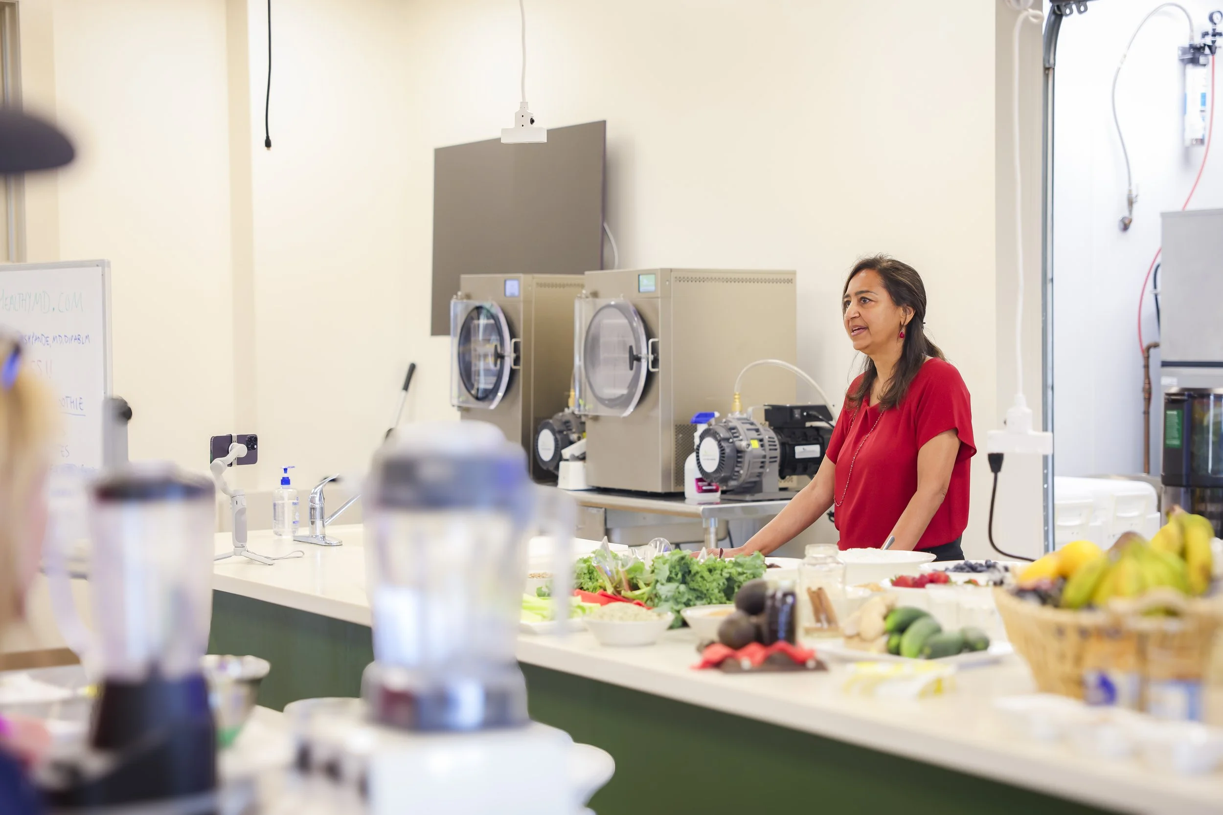 A woman in a red shirt giving a cooking demonstration at a kitchen counter covered with fresh vegetables and kitchen appliances.