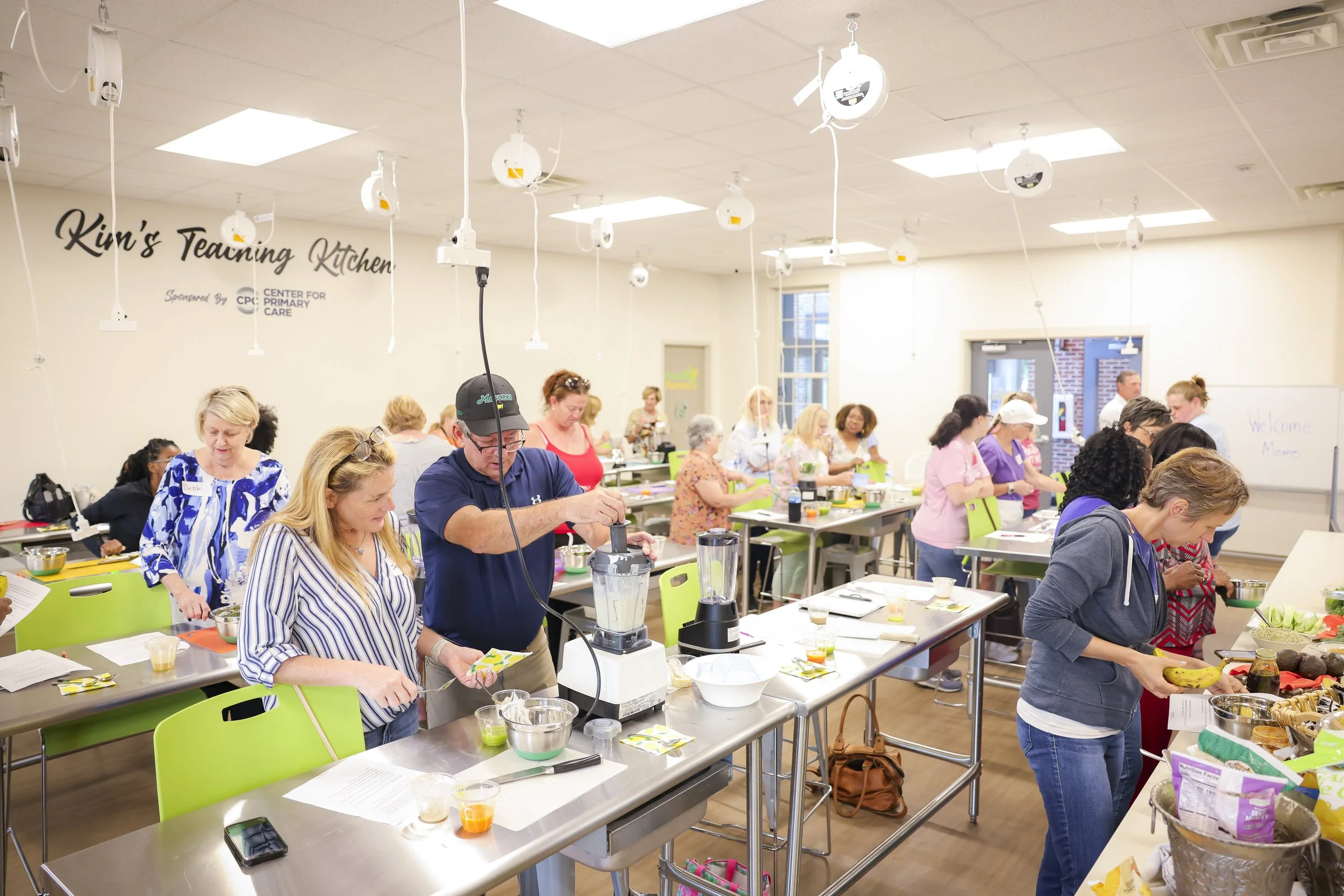Women and men participating in a cooking class in a brightly lit kitchen classroom, with various ingredients and cooking tools on the tables, some preparing food.