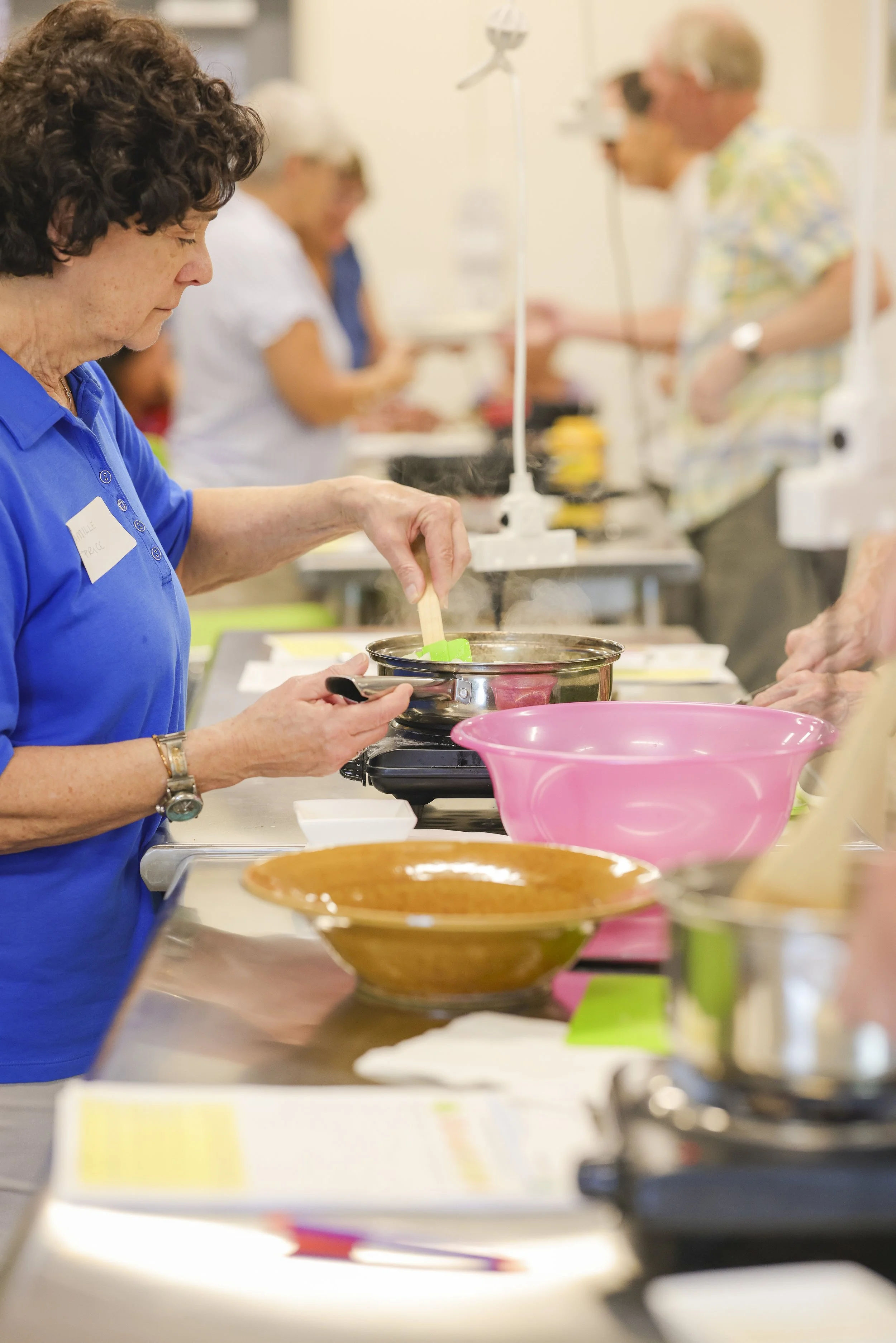 Women participating in a cooking class, stirring ingredients on a stove with various cooking utensils and bowls on the counter.