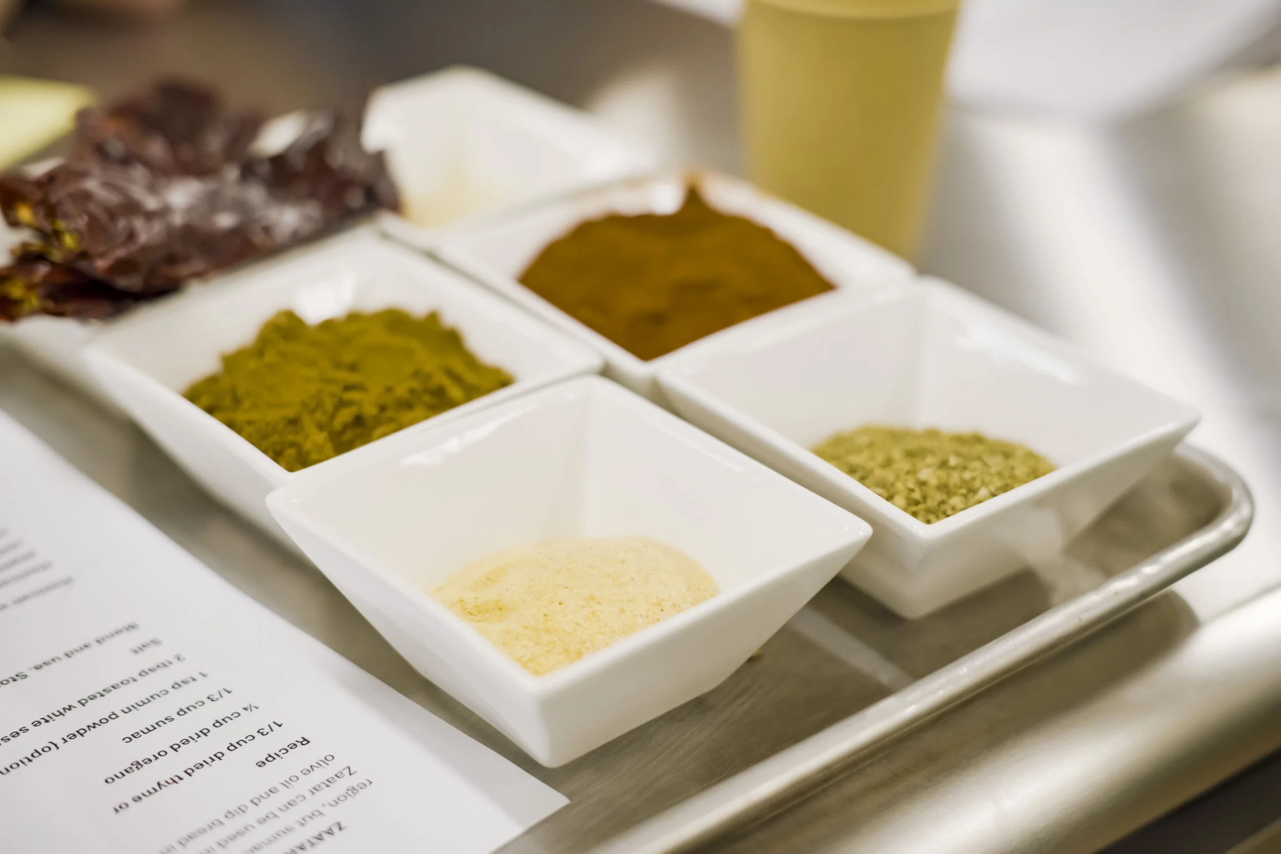 Six white square dishes containing various spices and herbs, placed on a tray on a table with a menu and a glass in the background.