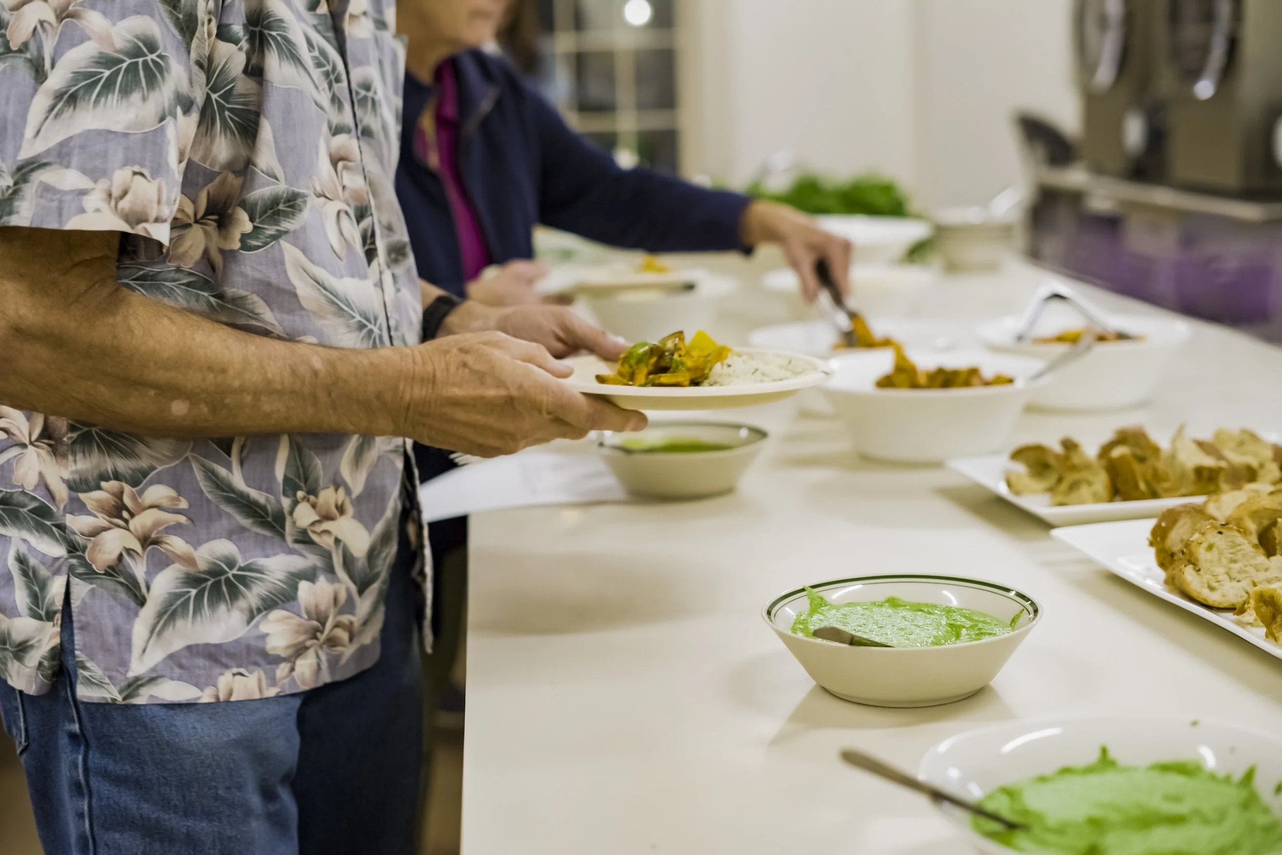 People serving themselves food at a buffet, with dishes including green sauce, fried items, and rice.