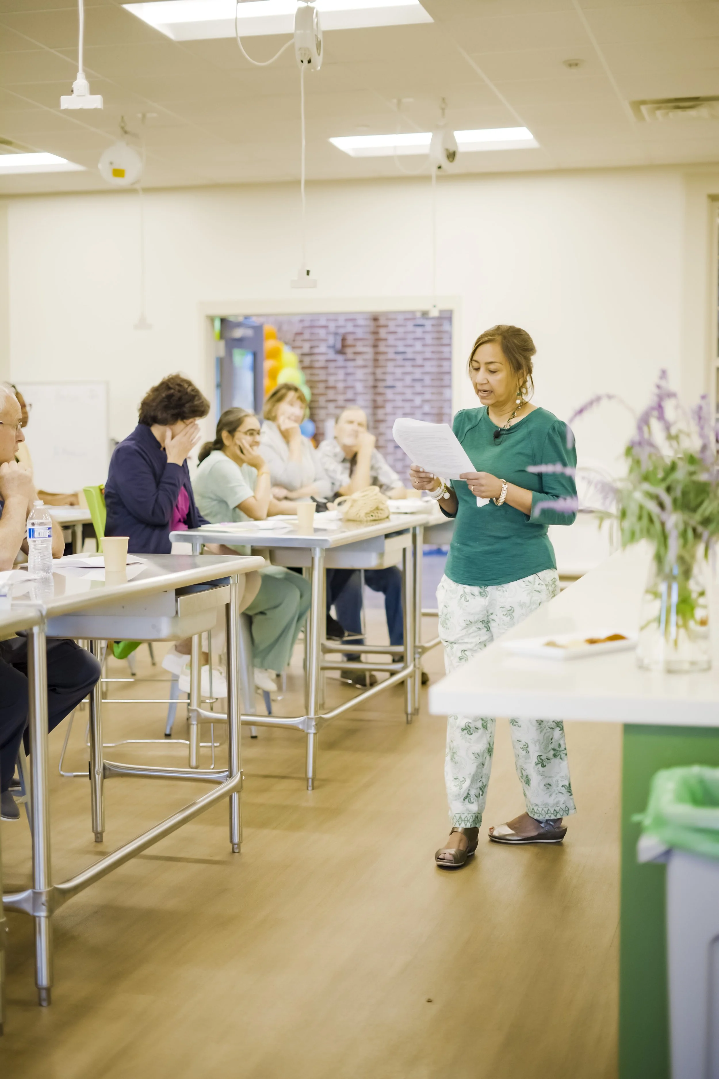 A woman standing and reading a paper to a seated group of people in a classroom or workshop setting.