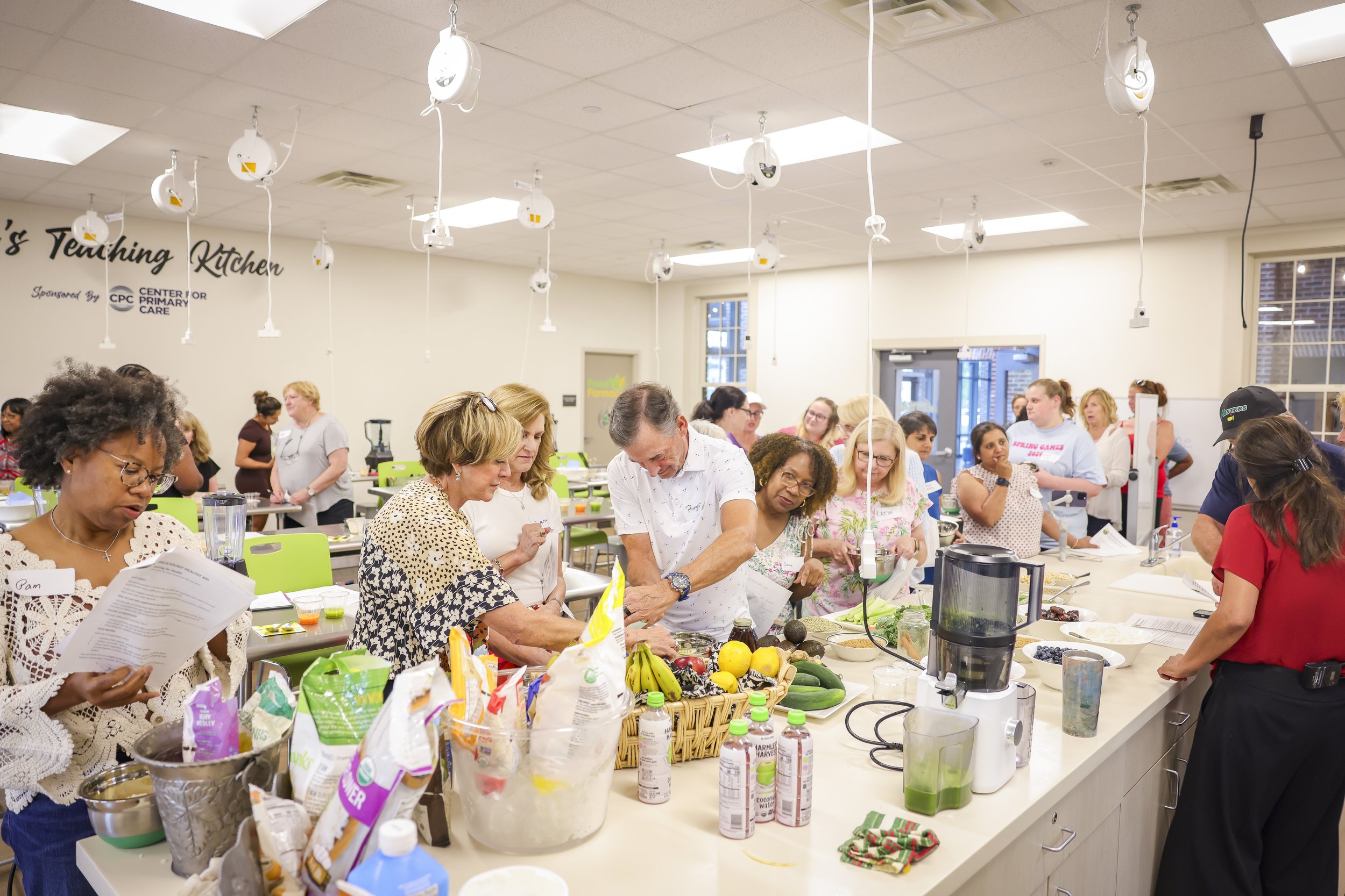 People participating in a cooking class in a kitchen with a white countertop, various fresh fruits and vegetables, and kitchen appliances.