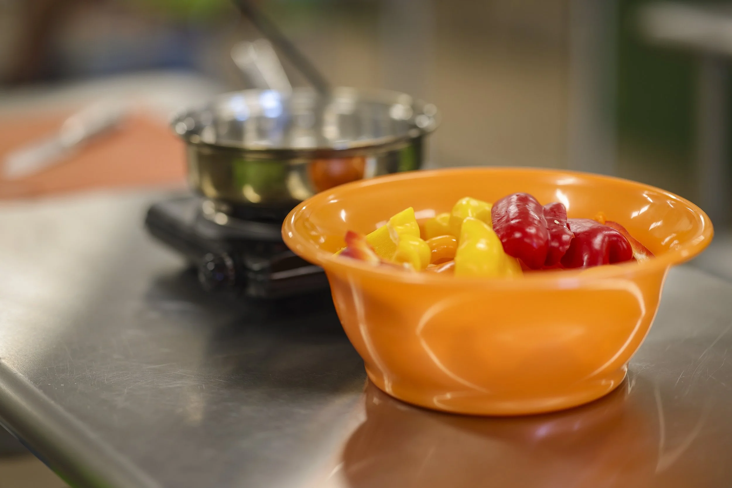 Orange bowl filled with mixed red and yellow bell peppers on a stainless steel countertop, with a small stainless steel pot and stove burner in the background.