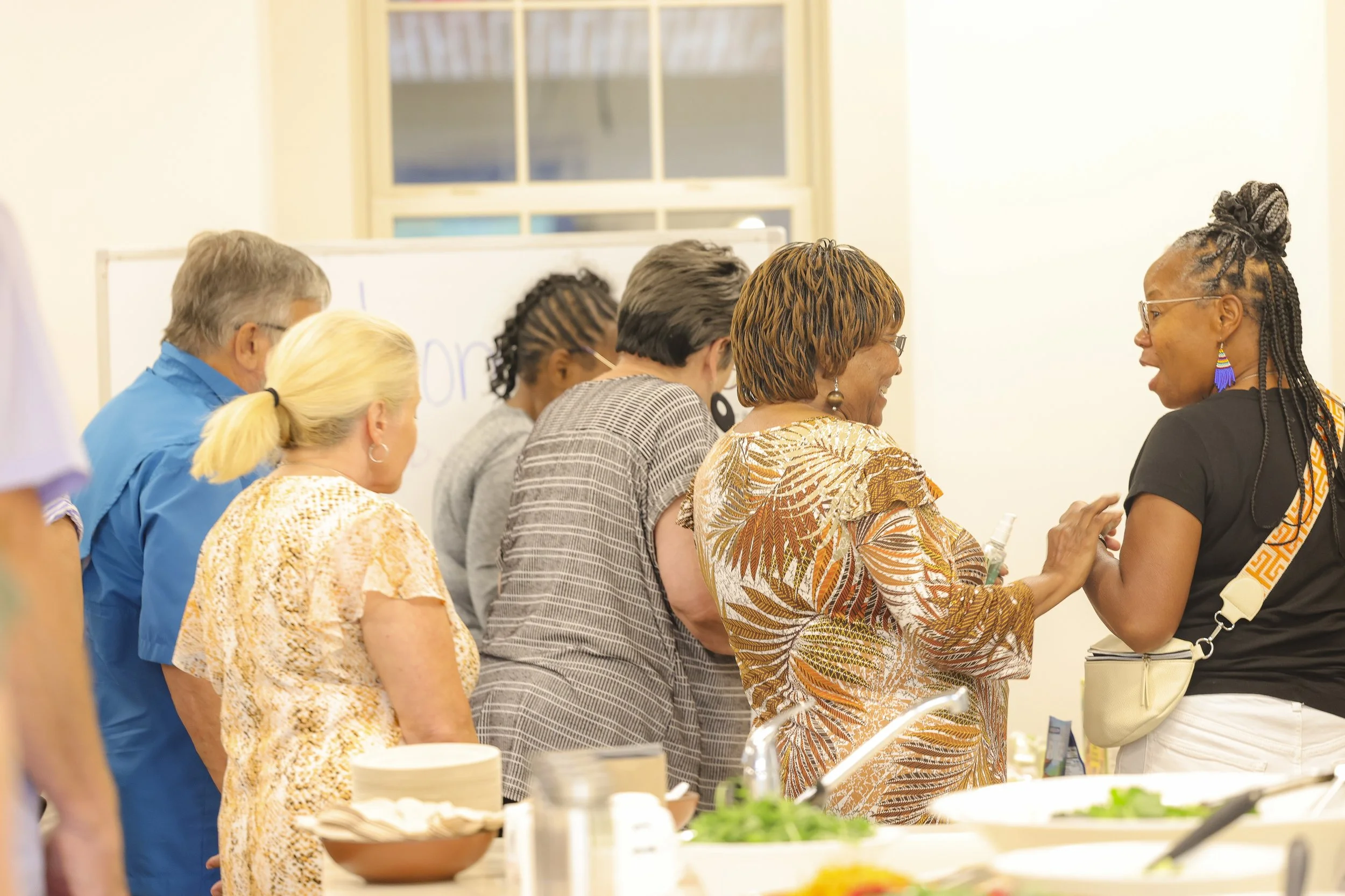 A group of six diverse adults line up at a buffet table, engaging in conversation and laughing, in a well-lit indoor setting.