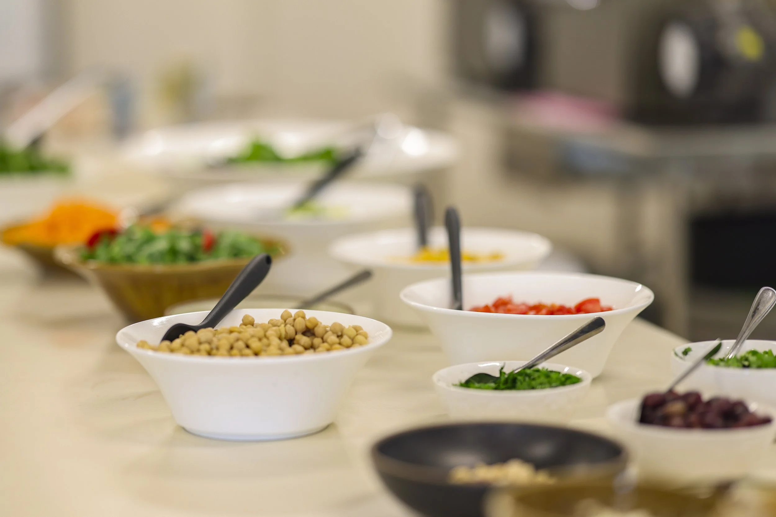 A variety of salad ingredients in white bowls on a table, including chickpeas, chopped tomatoes, greens, nuts, and other vegetables.